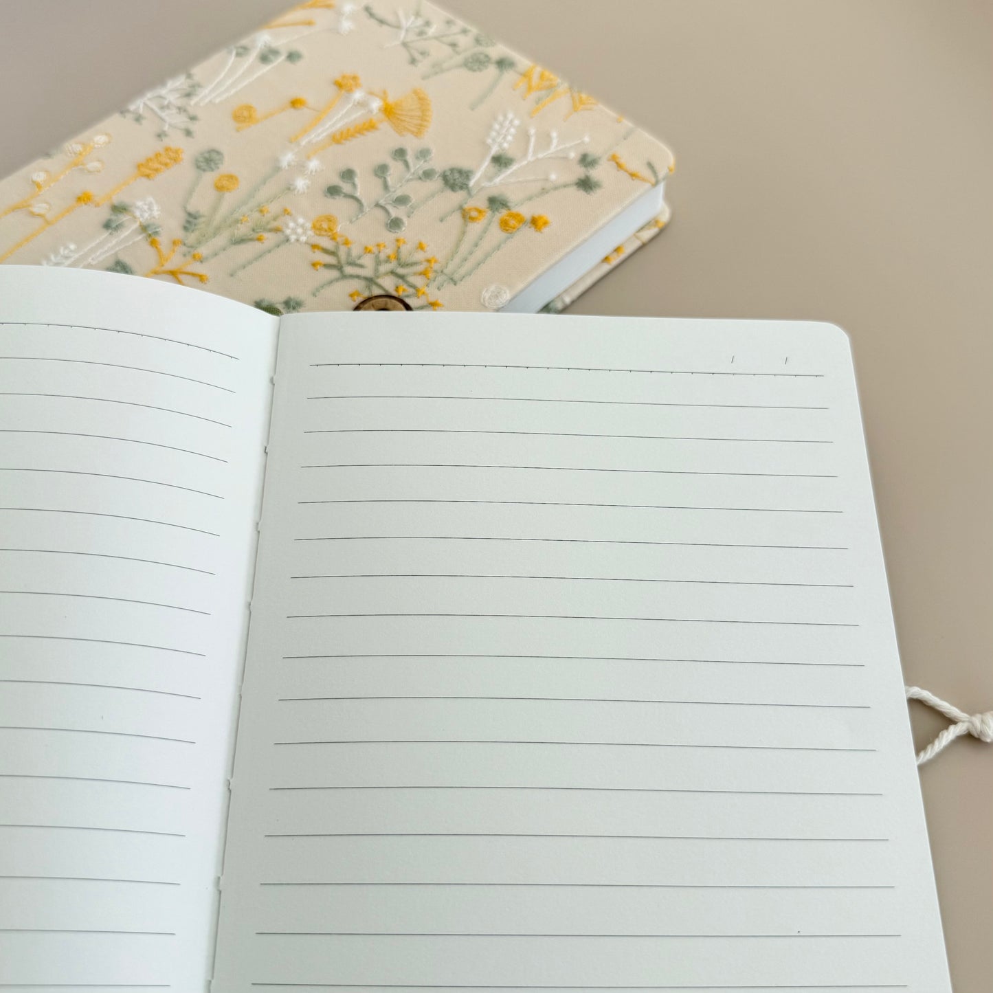 An open lined notebook with blank white pages in the foreground and a closed floral-patterned notebook in the background on a neutral beige surface.