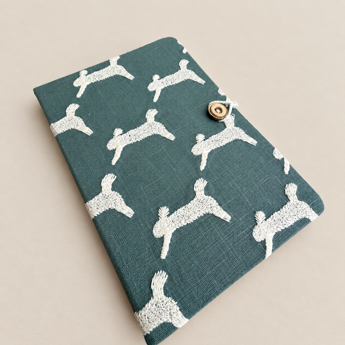 A teal fabric notebook or journal cover with a white embroidered dog pattern and a small gold button closure, shown on a neutral beige background.