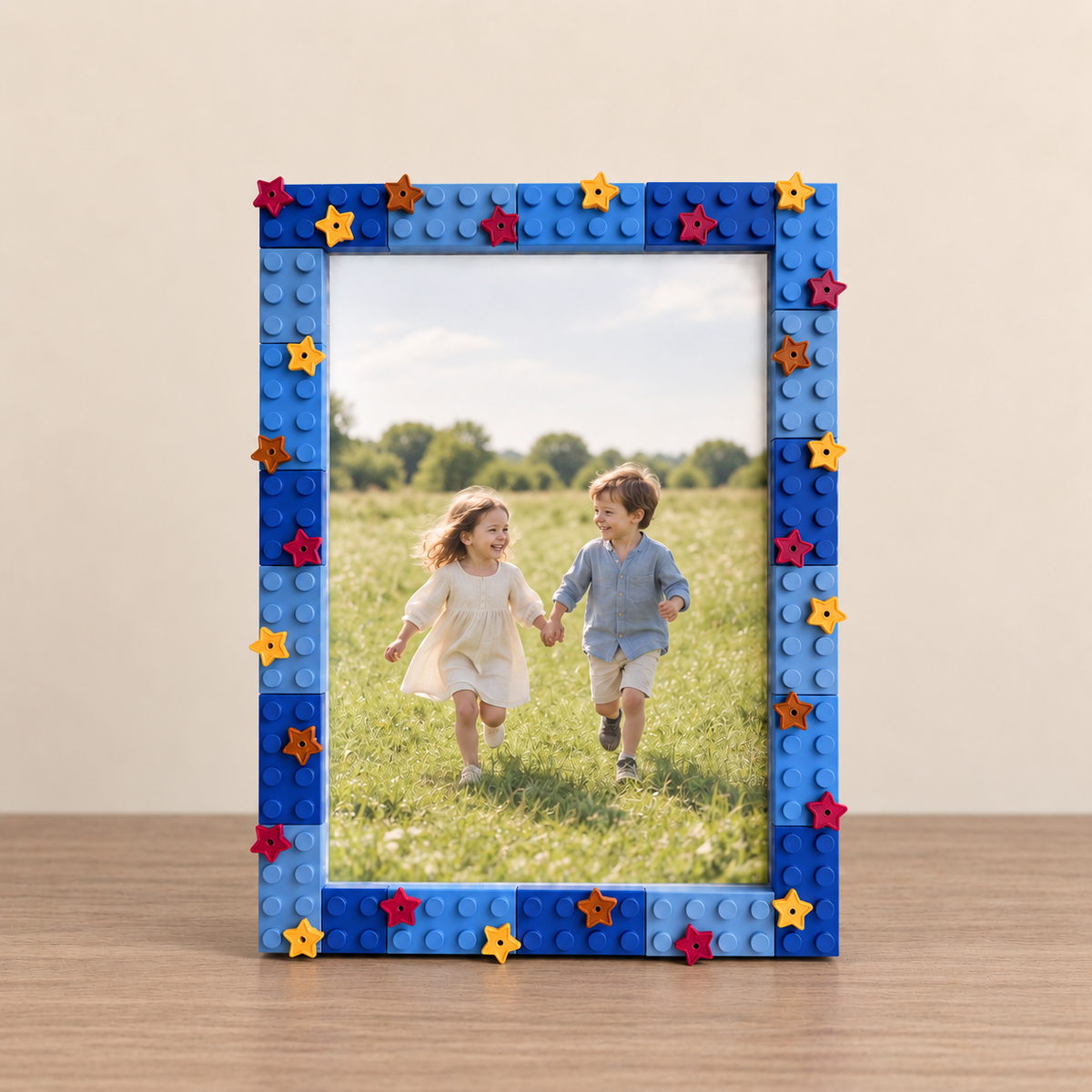 A square photo frame made from blue building bricks with small star-shaped studs in red, orange, and yellow, holding a photo of two children running in a grassy field. The frame sits on a wooden surface against a plain beige wall.