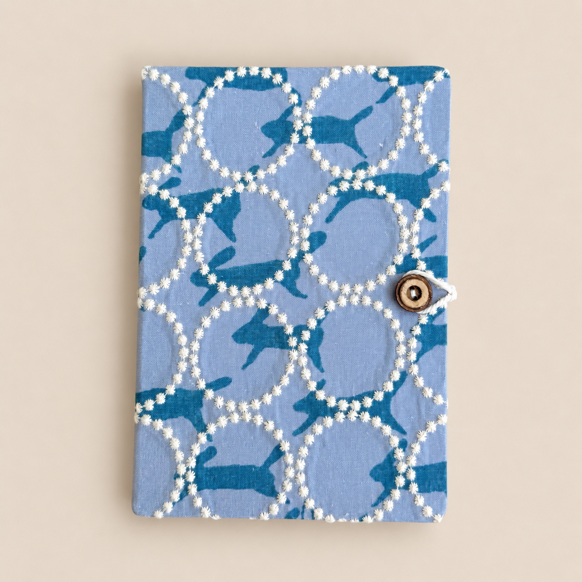 A notebook with a light blue cover featuring a pattern of dark blue silhouettes and white beaded circles, secured with a small wooden button closure, set against a solid beige background.