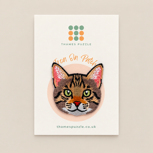 An embroidered cat patch displayed on its branded backing card. The patch features a tabby cat's face with green eyes, pink inner ears, and a small orange nose.