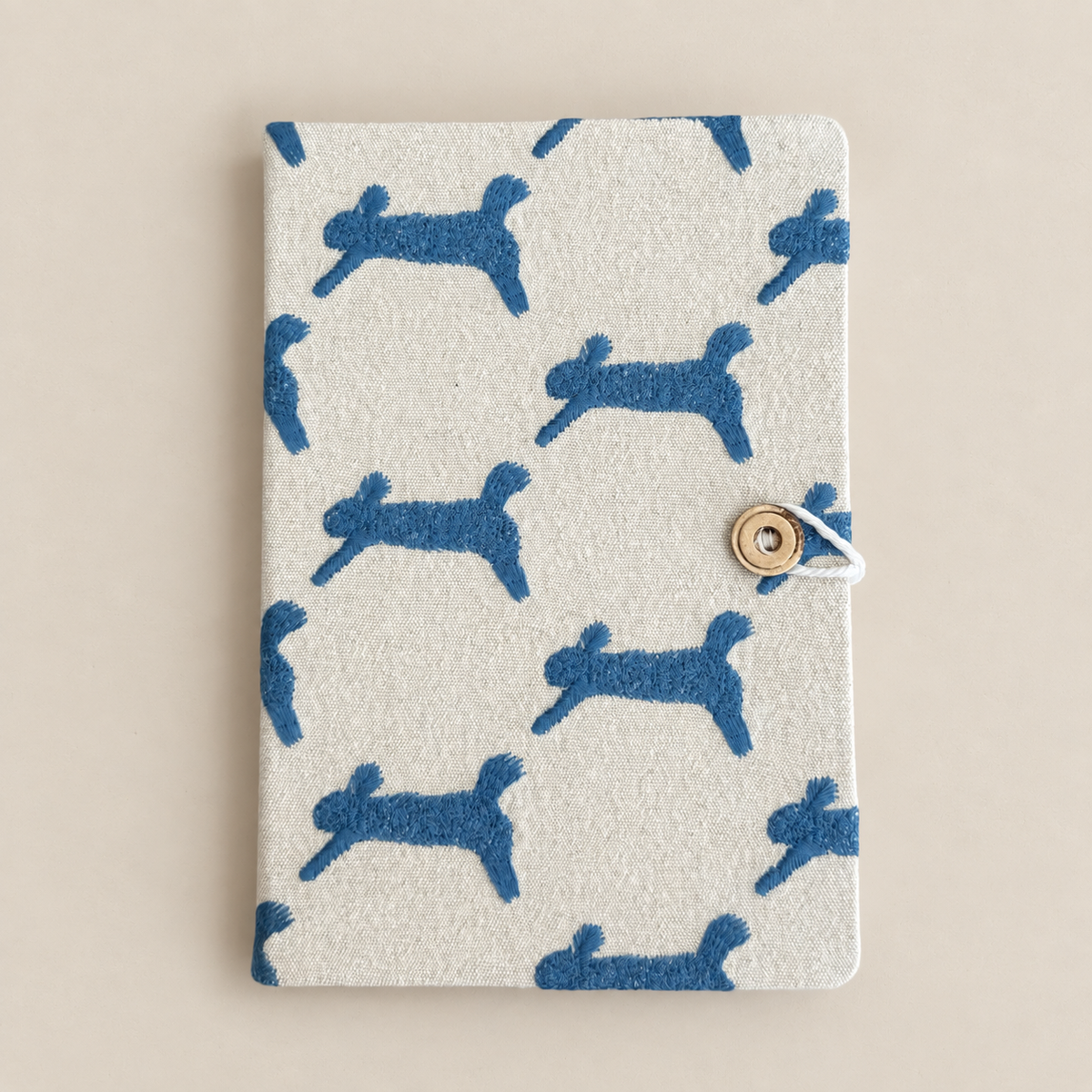 A rectangular fabric notebook or journal with a light beige cover featuring a repeating blue silhouette pattern of dogs, secured with a small metal snap button and white cord on the right side, photographed on a plain beige background.