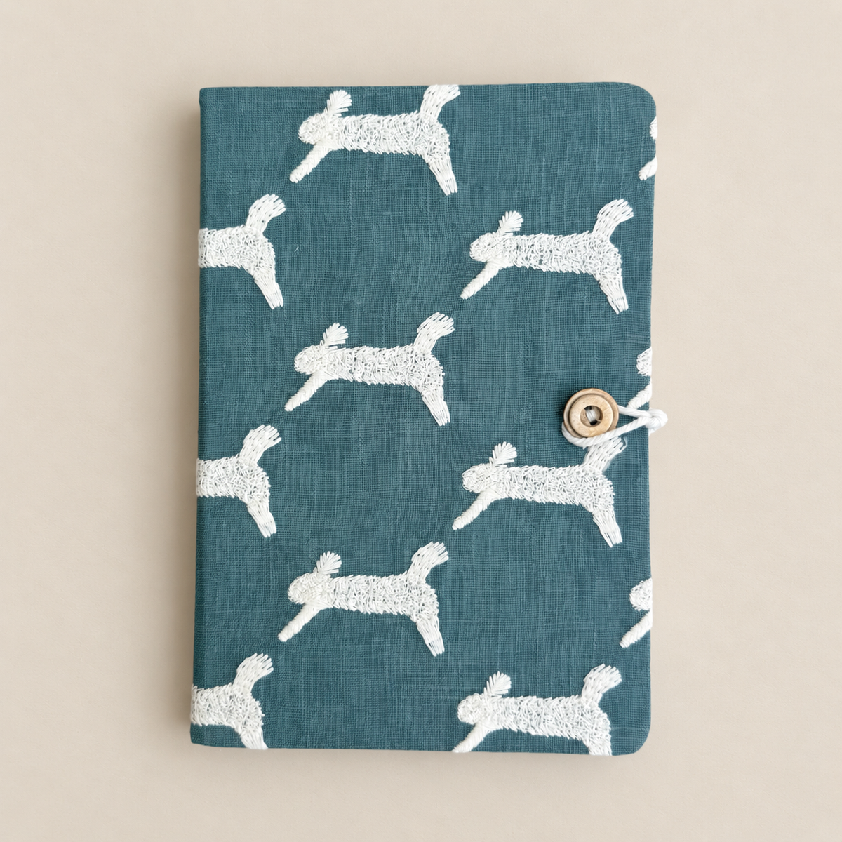 A fabric-covered notebook or journal with a teal blue-green background and a repeating white dog silhouette pattern, secured with a small metal snap button and white cord on a warm beige background.