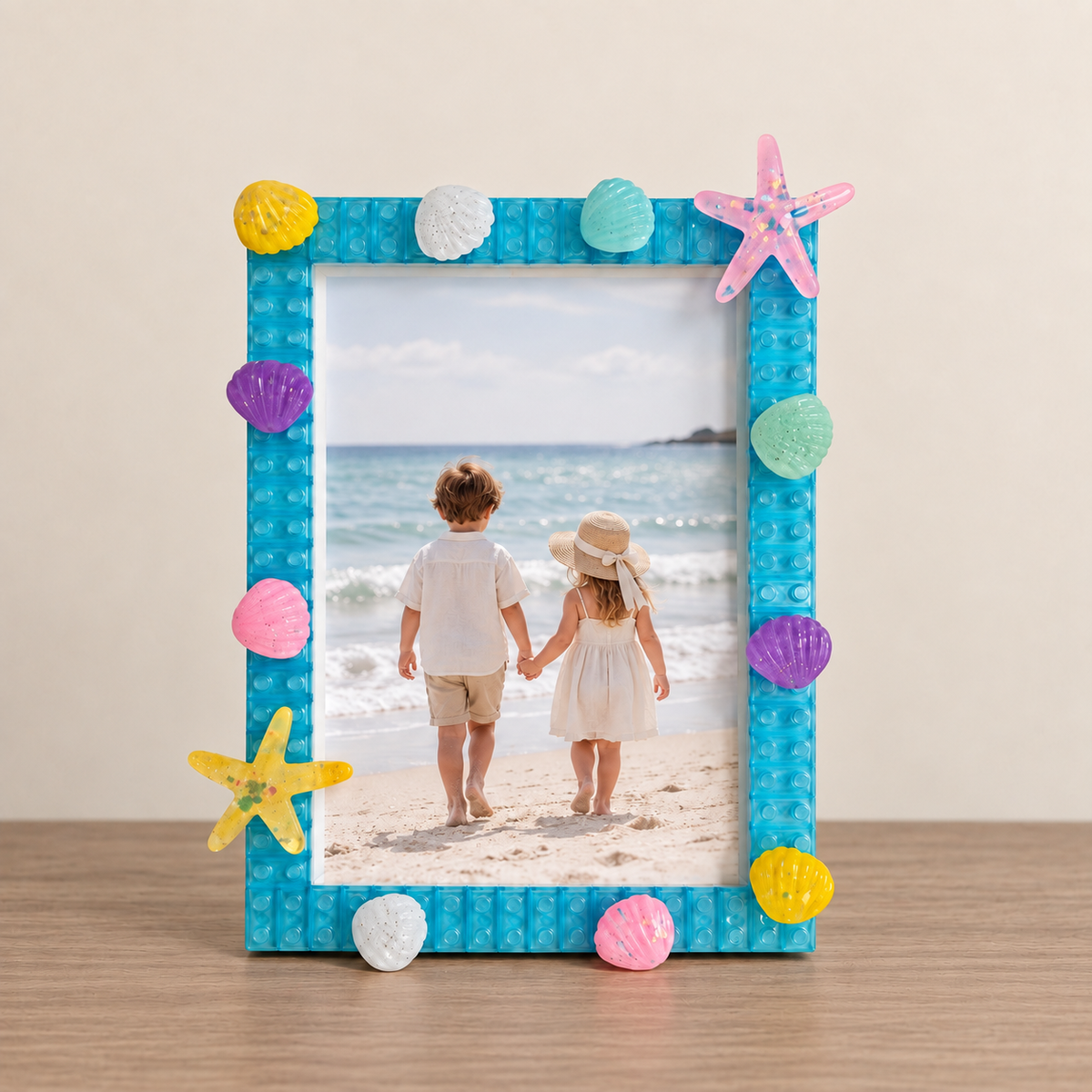 A square photo frame with a bright blue brick-like border, decorated with colorful seashells and starfish accents, holding a photo of two children on a beach, set on a wooden surface against a beige wall.