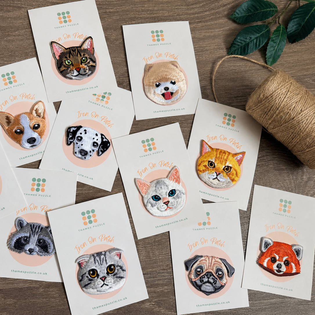 A styled flat-lay of multiple custom-printed cards or stickers featuring illustrated cat faces, arranged on a wooden surface with a spool of twine and green leaves nearby. The cards show the text 'Just One More' and a small logo at the top.
