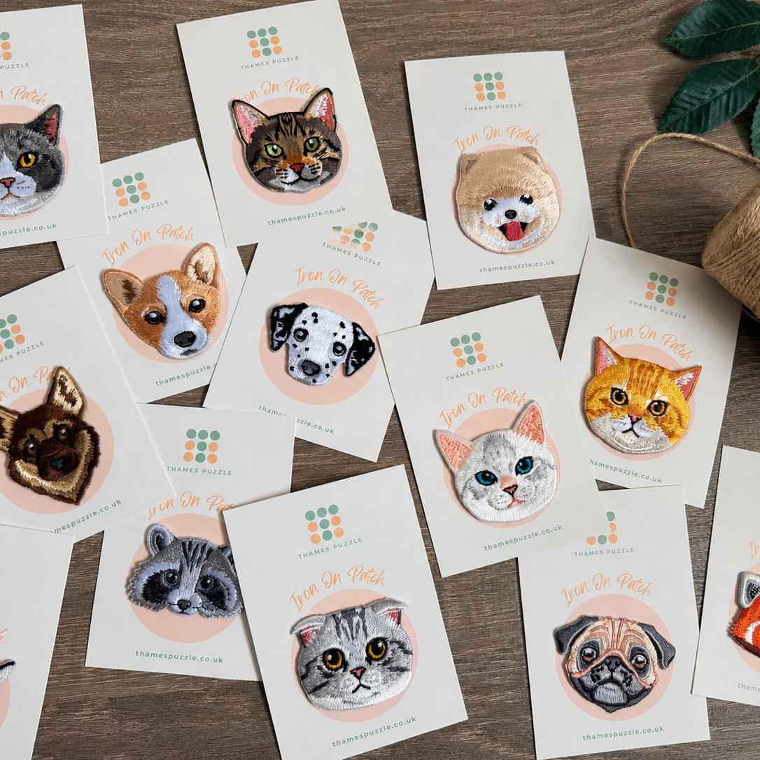 A styled flat-lay of multiple custom-printed cards or stickers featuring illustrated cat faces, arranged on a wooden surface with a spool of twine and green leaves nearby. The cards show the text 'Just One More' and a small logo at the top.
