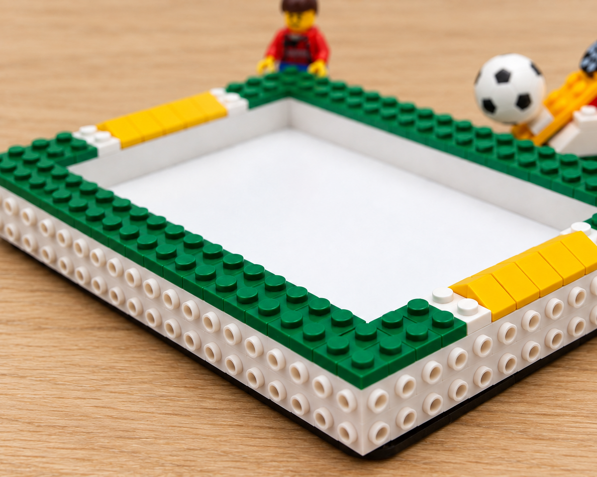 A brick  rectangular stadium or field frame made of green, white, and yellow bricks, photographed on a light wood surface with small toy minifigures and a soccer ball near the top edge.