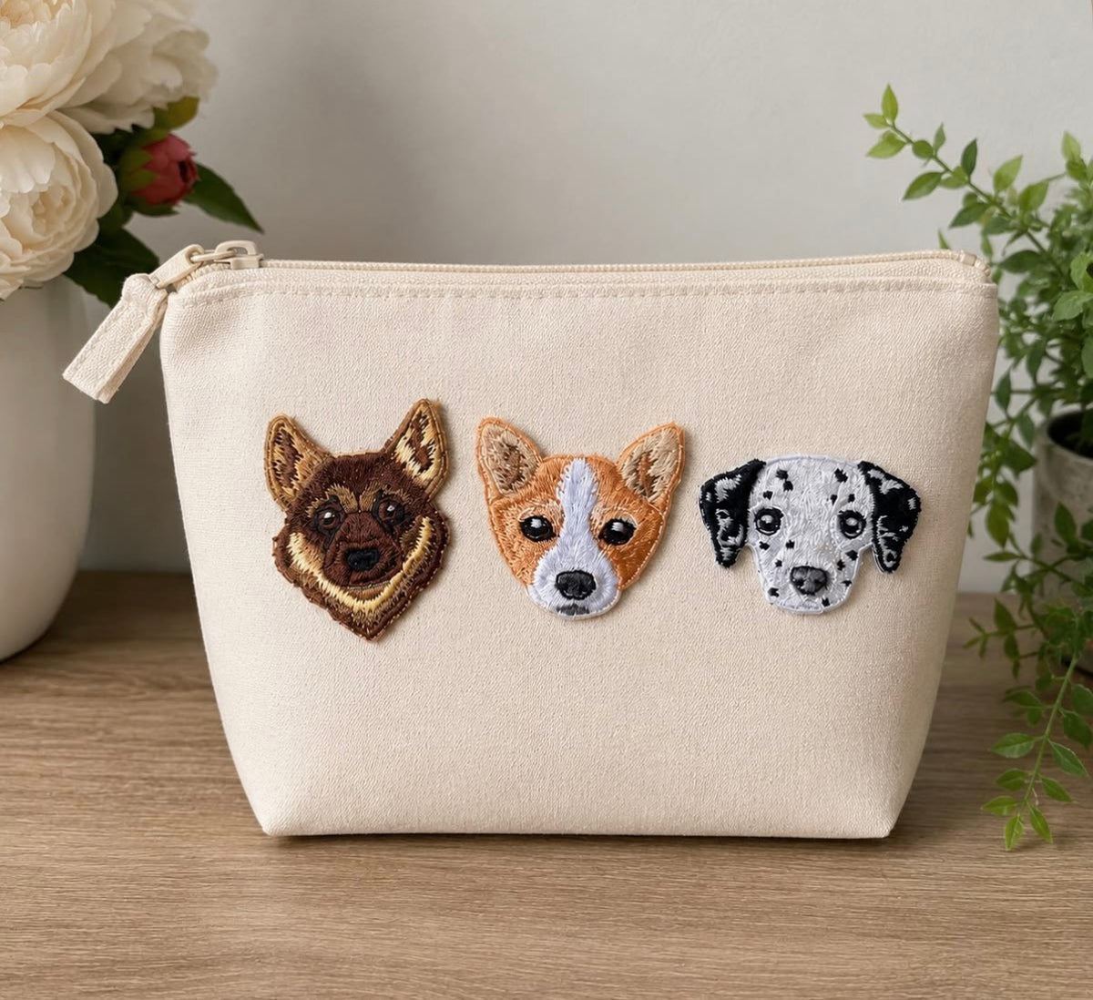 An off-white canvas zippered pouch with three embroidered dog faces (German Shepherd, Corgi, and Dalmatian) on the front, resting on a wooden surface with a blurred background of flowers and a plant.