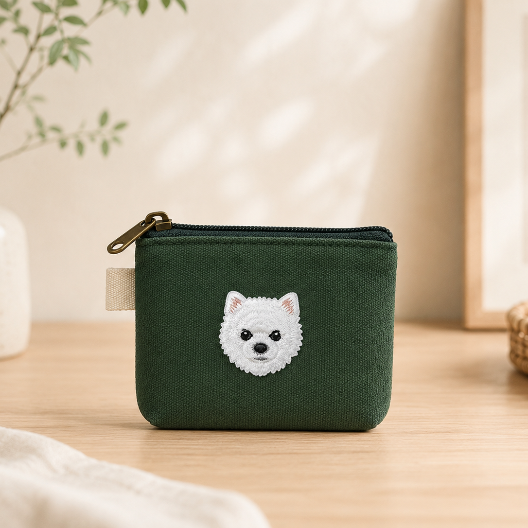 A dark green fabric zippered pouch with a white embroidered dog face on the front, sitting on a light wooden table in a softly styled room setting.