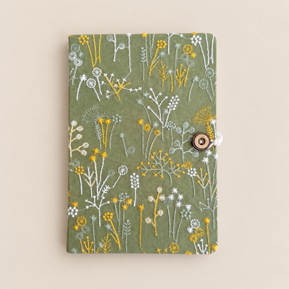 A fabric-covered notebook or journal with a muted sage green cover, decorated with a delicate white and mustard yellow wildflower pattern, and secured with a small wooden toggle closure on the right side.