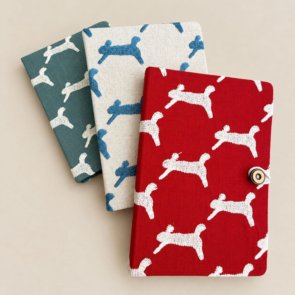 Three fabric-covered notebooks with a dog pattern, arranged diagonally on a light beige background. The notebook in front is red with white dog silhouettes and a metal snap button closure, while the other two are teal and off-white respectively.