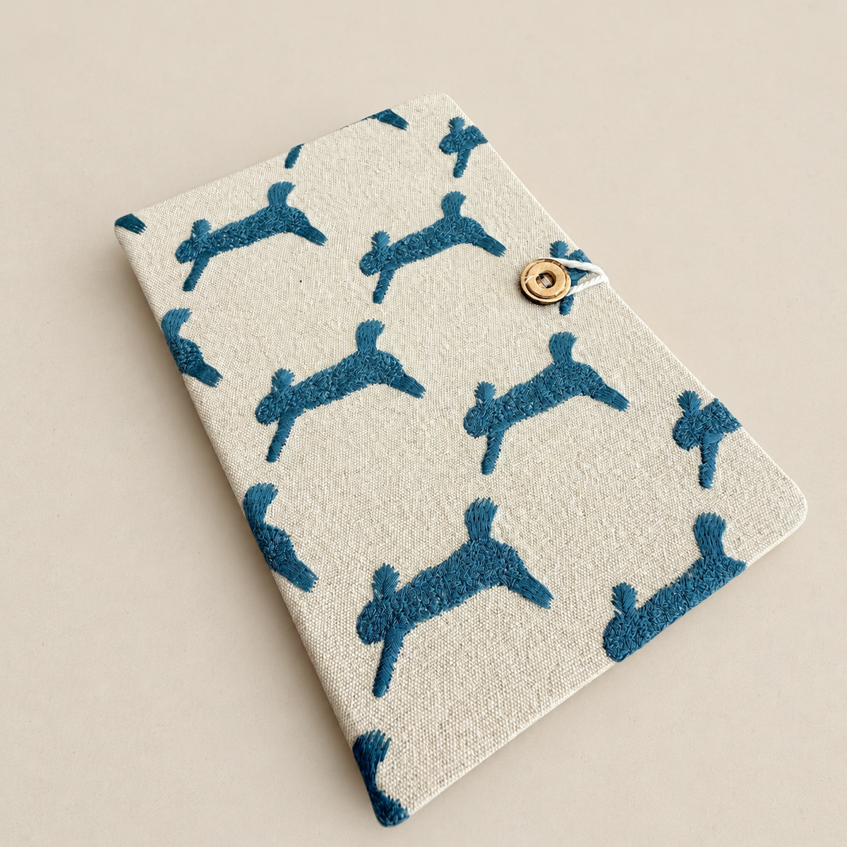 A rectangular fabric notebook or journal cover with a cream linen-like background and repeating dark blue dog silhouettes, secured by a small gold-tone snap button closure, photographed on a plain warm beige background.