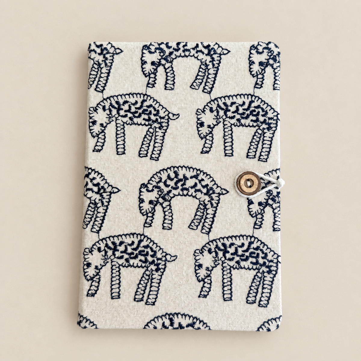A fabric notebook or journal cover featuring a repeating navy blue hand-drawn elephant pattern on a warm off-white background, secured with a small round wooden toggle clasp on the right side.