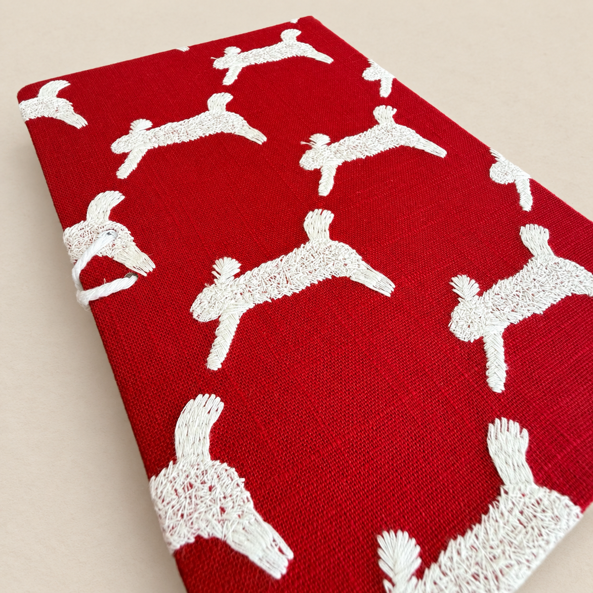 A close-up of a red woven fabric with a repeating white embroidered dog pattern, laid flat on a light beige background.