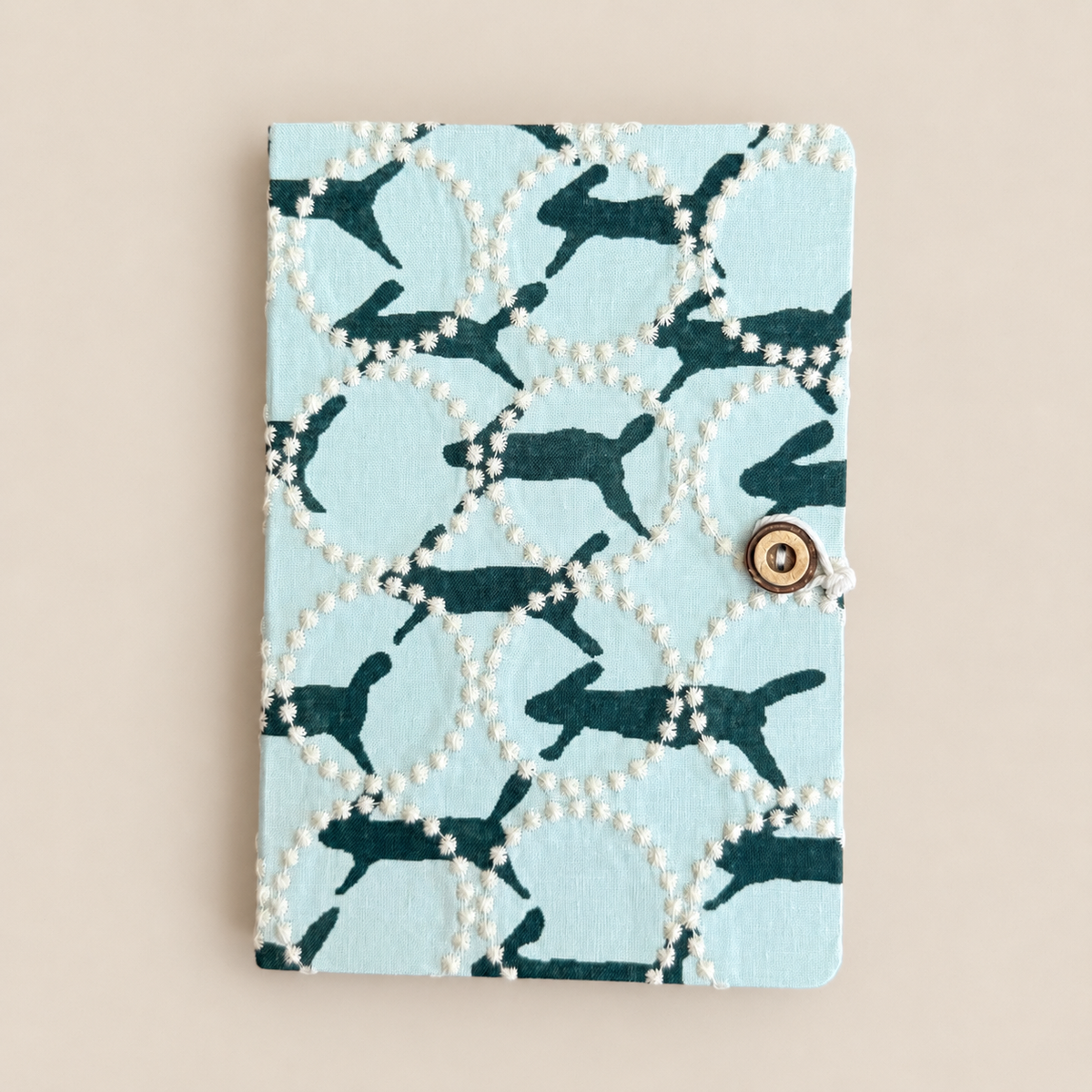 A square notebook or journal with a light blue cover featuring a pattern of dark teal rabbits inside white circular lattices outlined with small white pearls. It has a small gold snap button closure on the right side and is photographed flat on a warm beige background.