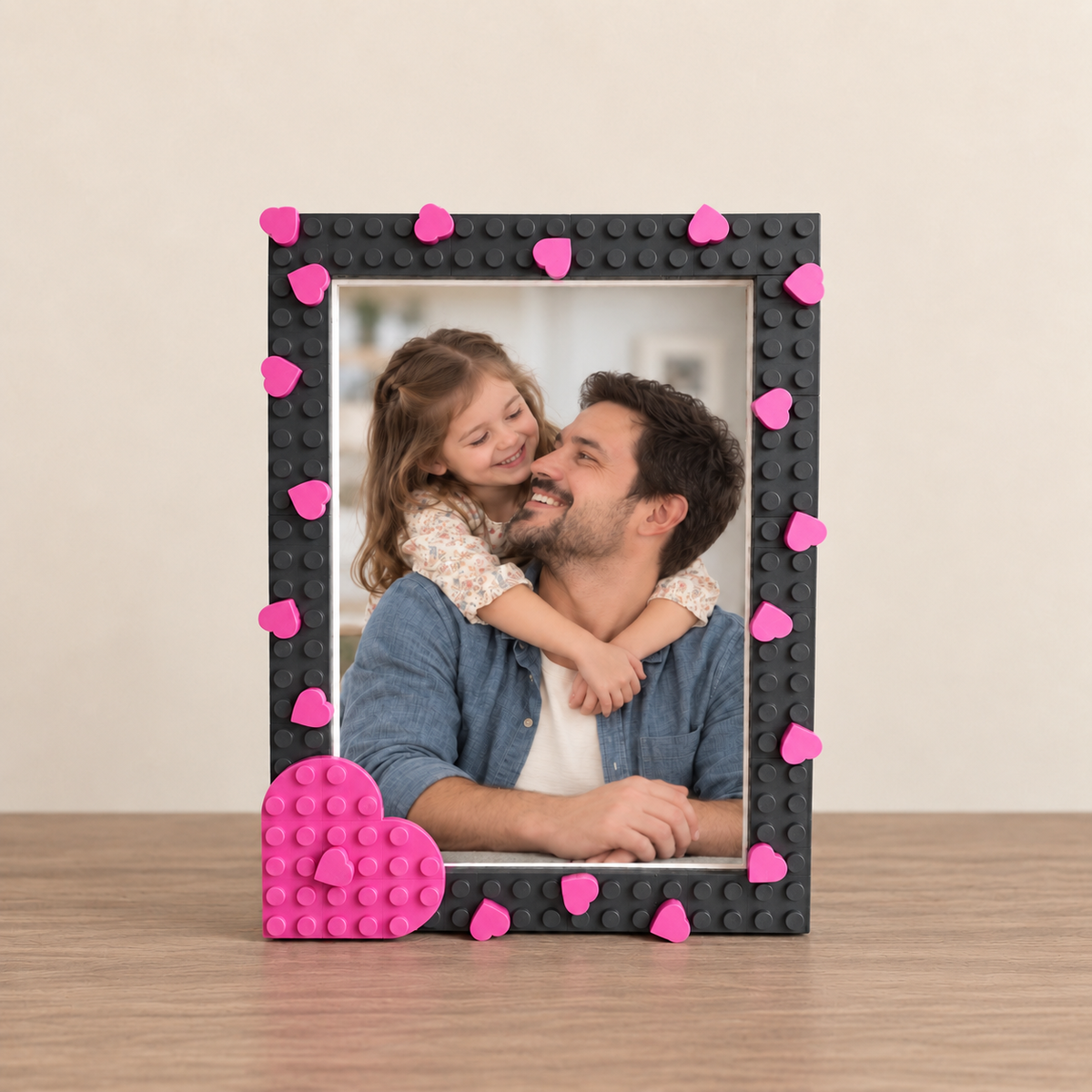 A black picture frame with bright pink heart studs and a matching pink heart base, holding a photo of a man and child, set on a wooden surface against a beige wall.