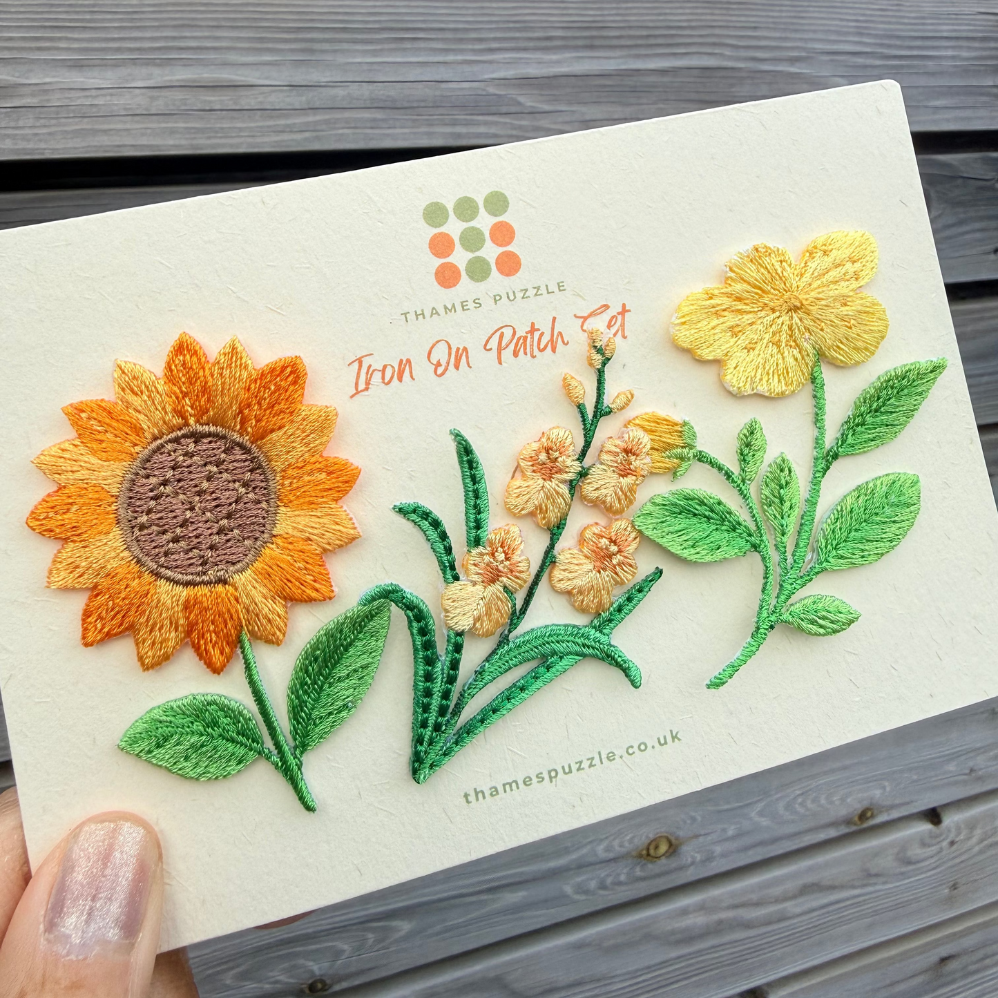 A hand holds a cream-colored iron-on patch card featuring embroidered flowers, including a large orange sunflower, yellow blossoms, and green leaves. The card has the 'Thames Puzzle' logo and branding printed on it, set against a gray wooden plank background.