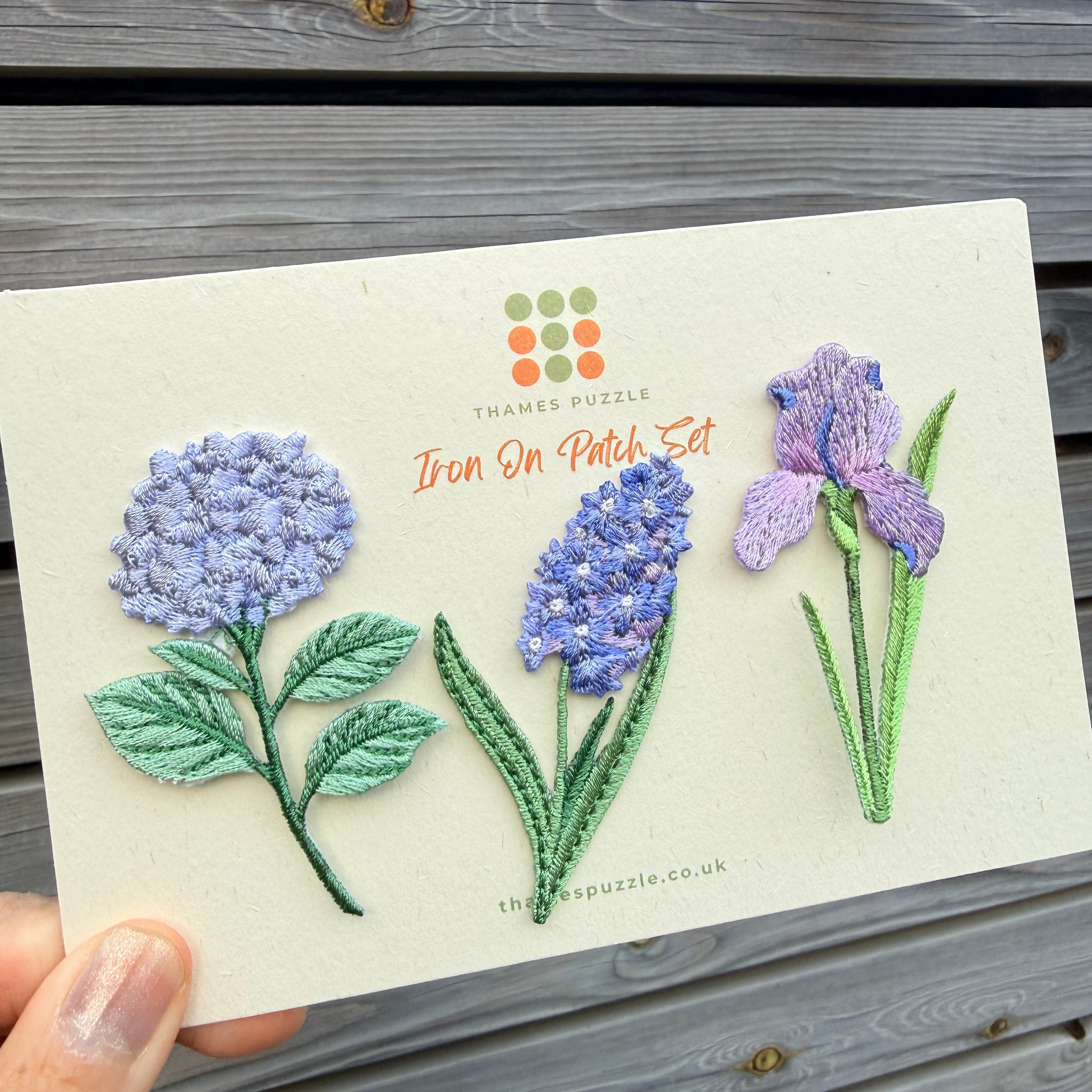 A hand holds a cream-colored card featuring three embroidered floral designs: a light purple hydrangea, a blue-purple hyacinth, and a lavender iris, with the text 'THAMES PUZZLE Iron On Patch Set' on the card, set against a gray wooden plank background.