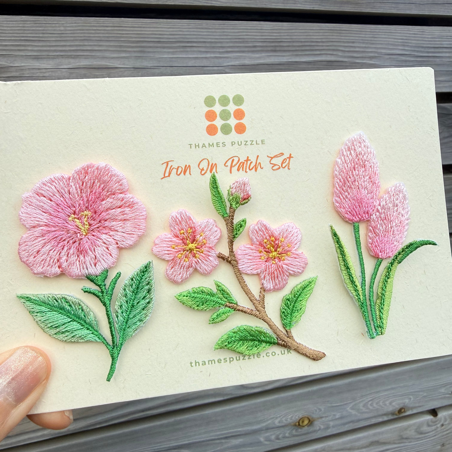 A hand holds a cream-colored card featuring embroidered floral designs, including pink flowers and green leaves, with the text 'Thames Puzzle Iron On Patch Set' printed on it, set against a gray wooden plank background.