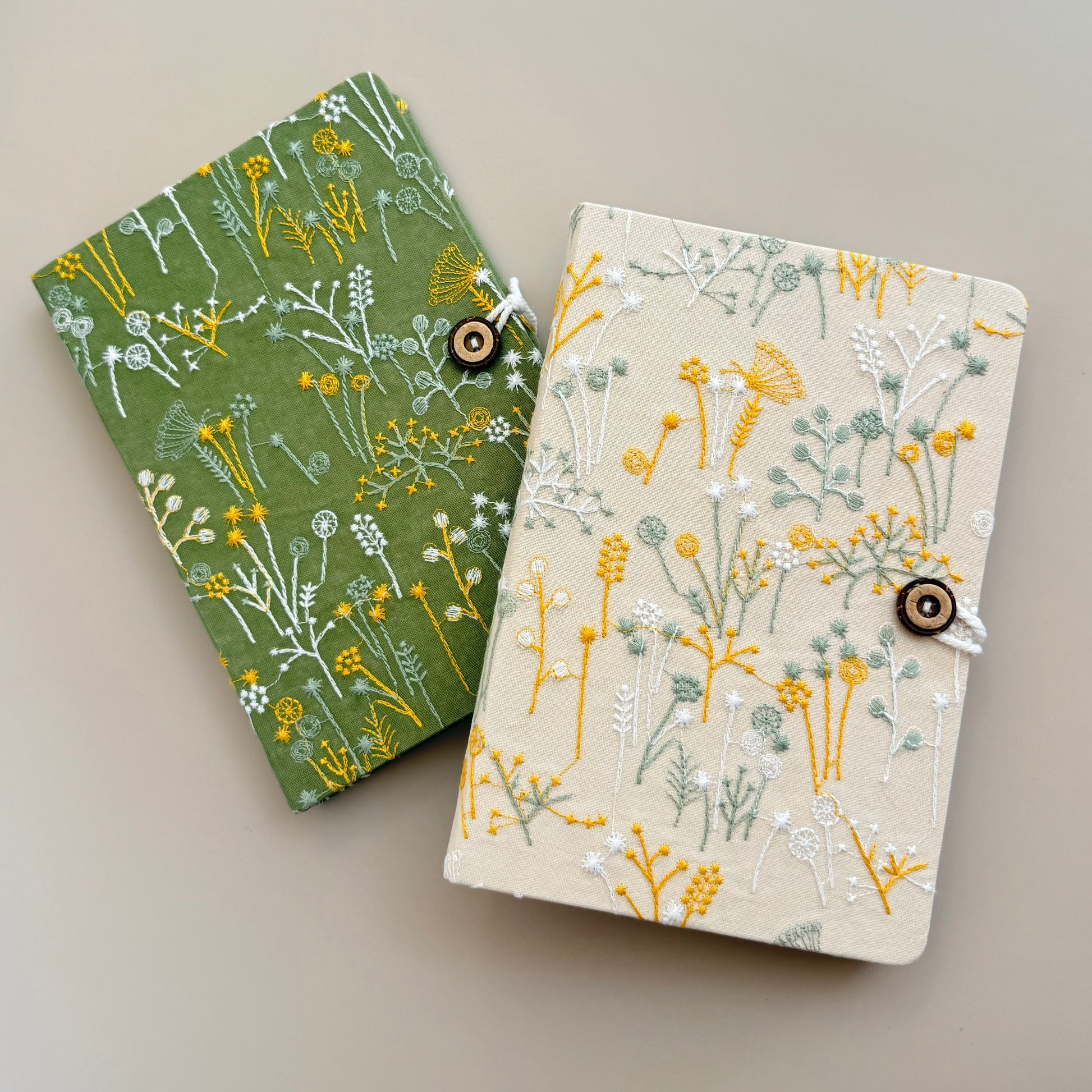 Two small fabric-covered notebooks or journal covers with a wildflower print, shown on a plain warm gray background. One cover is olive green with yellow and white floral motifs, and the other is cream with a similar floral print in yellow, white, and muted sage green.