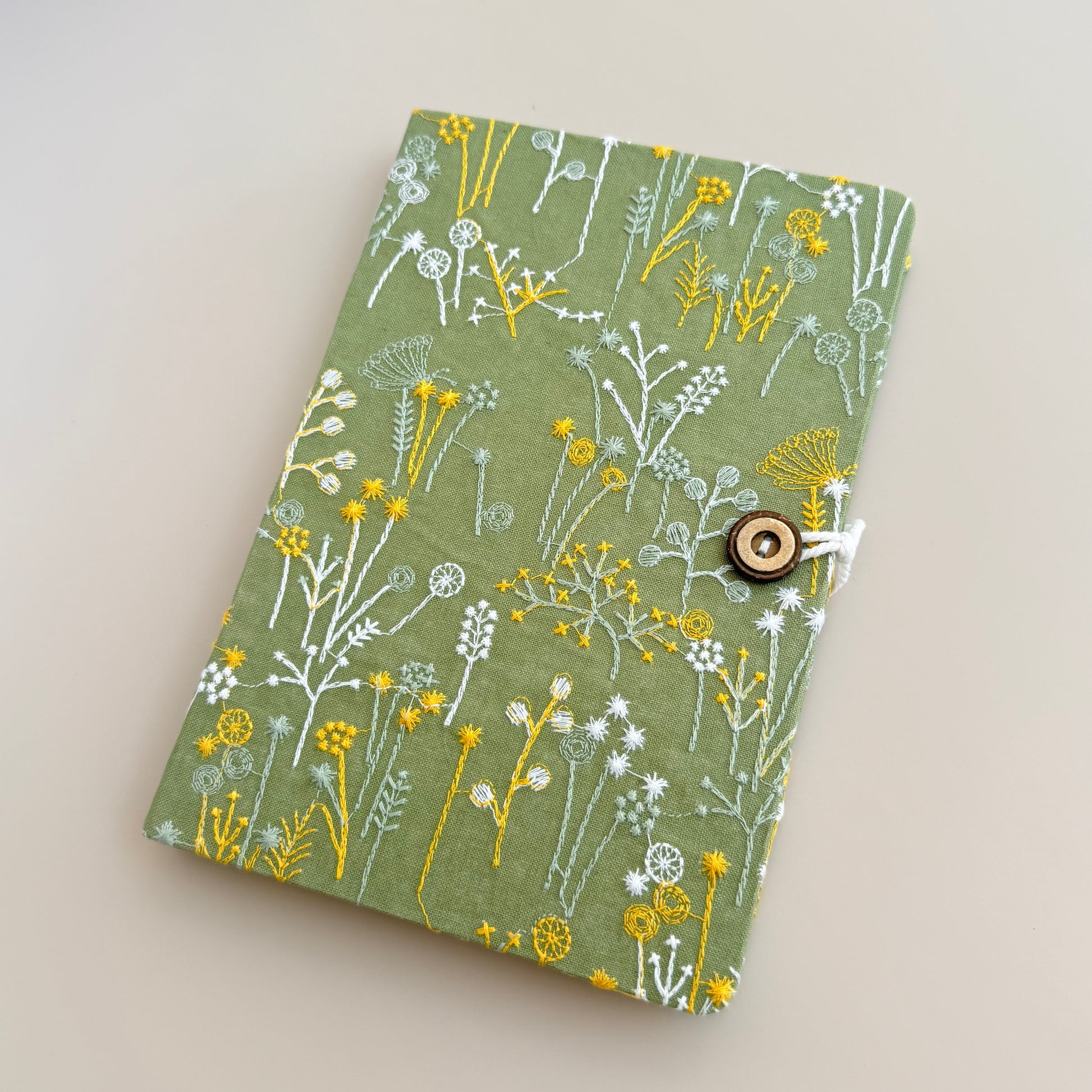 A fabric-covered notebook or journal with a green cover decorated with a white and yellow wildflower pattern, secured with a small wooden toggle clasp on a light beige background.