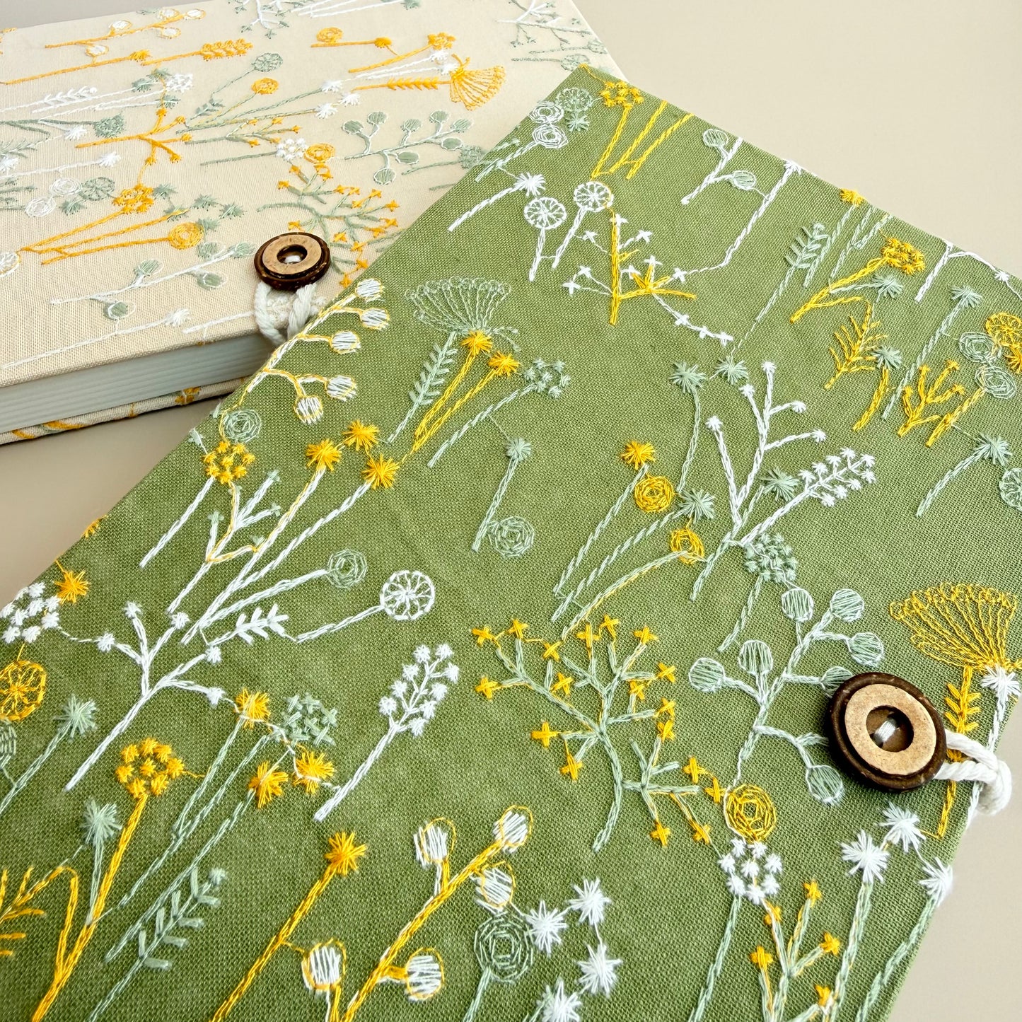 A close-up of a green fabric-covered notebook or journal with embroidered wildflowers in yellow and white, secured with a wooden button closure. A second notebook with a light beige cover and floral embroidery is partially visible in the background.