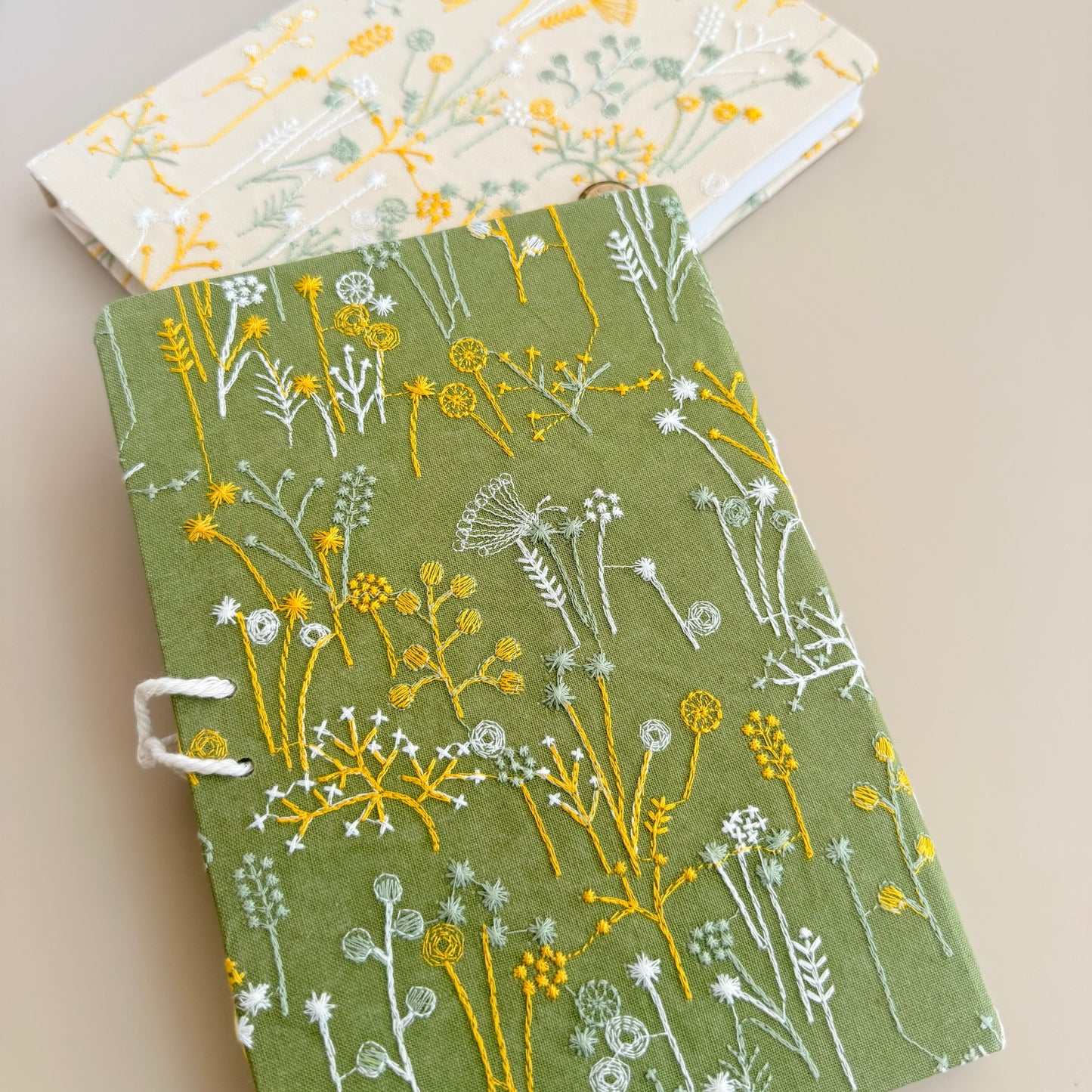 A hardcover journal with an olive green cover decorated with embroidered wildflowers in yellow and white, shown on a beige background with a second floral journal partially visible behind it.