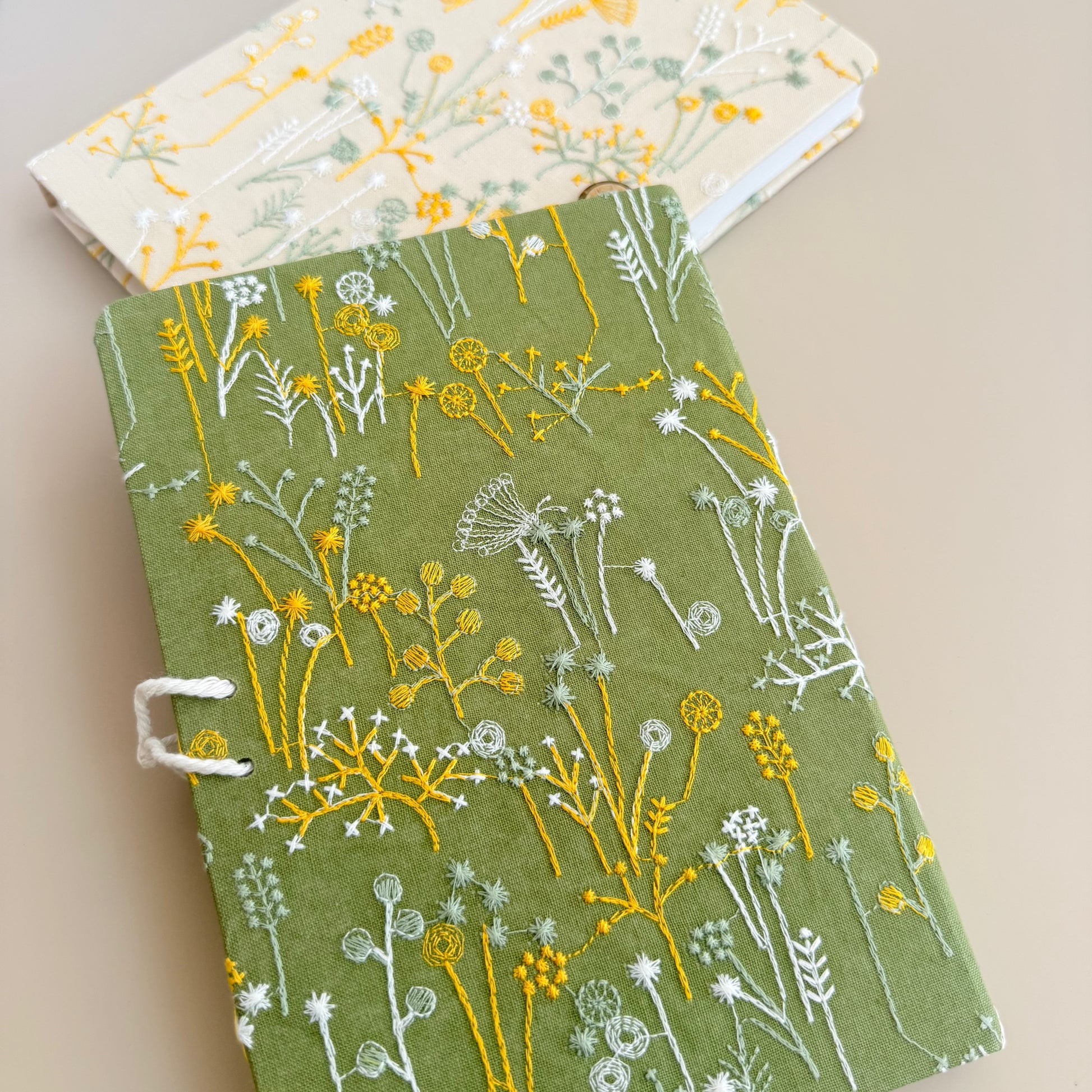 A hardcover journal with an olive green cover decorated with embroidered wildflowers in yellow and white, shown on a beige background with a second floral journal partially visible behind it.