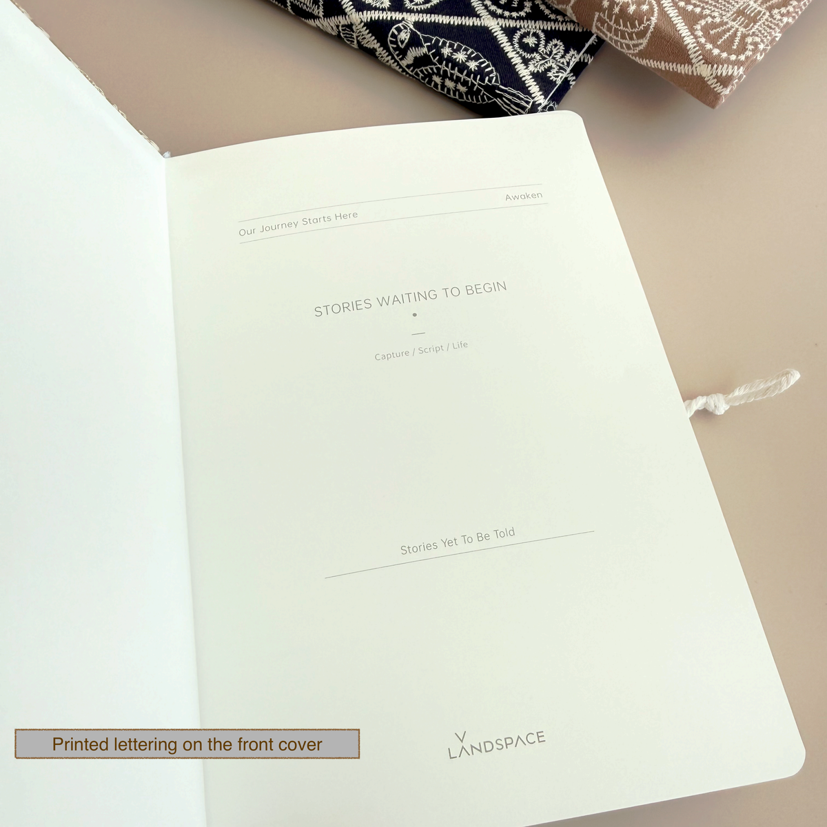 An open white notebook or journal laid on a beige surface, showing a printed interior page with placeholder text and the brand name 'V LANDSPACE' near the bottom. A small label at the lower left reads 'Printed lettering on the front cover.
