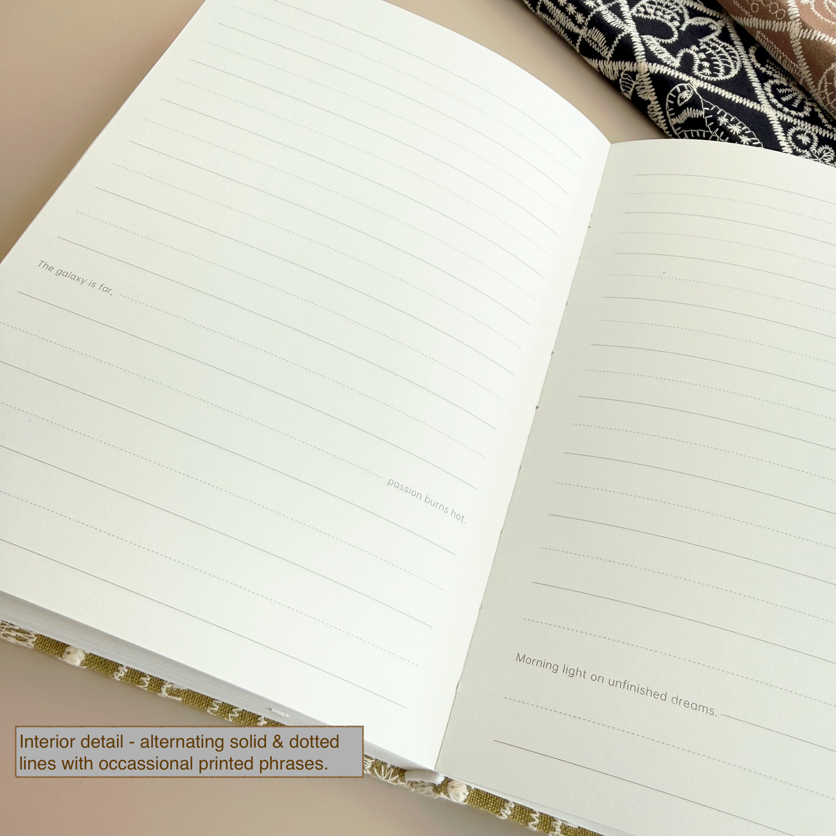 An open notebook showing lined interior pages with printed phrases, photographed on a beige surface with a patterned fabric partially visible at the top right.