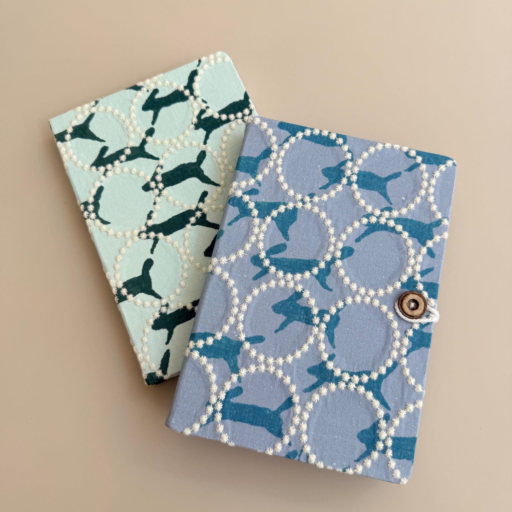 Two patterned fabric-covered notebooks or journal covers arranged on a plain beige background. The front cover is light blue with a teal dachshund print outlined by white pearl-like circles and a small metal snap closure.