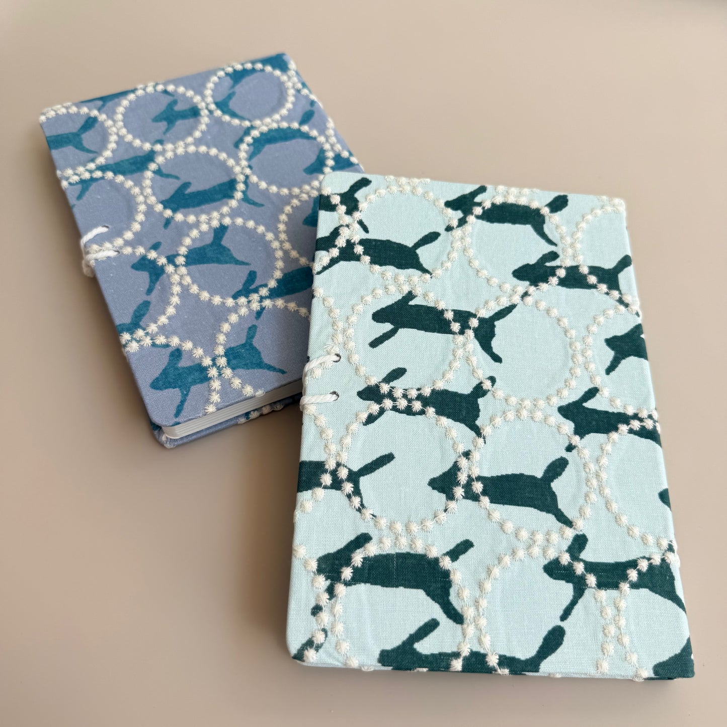 Two decorative notebooks with white pearl-bead borders and printed animal motifs, one in lavender-blue and one in pale blue, displayed on a beige background.