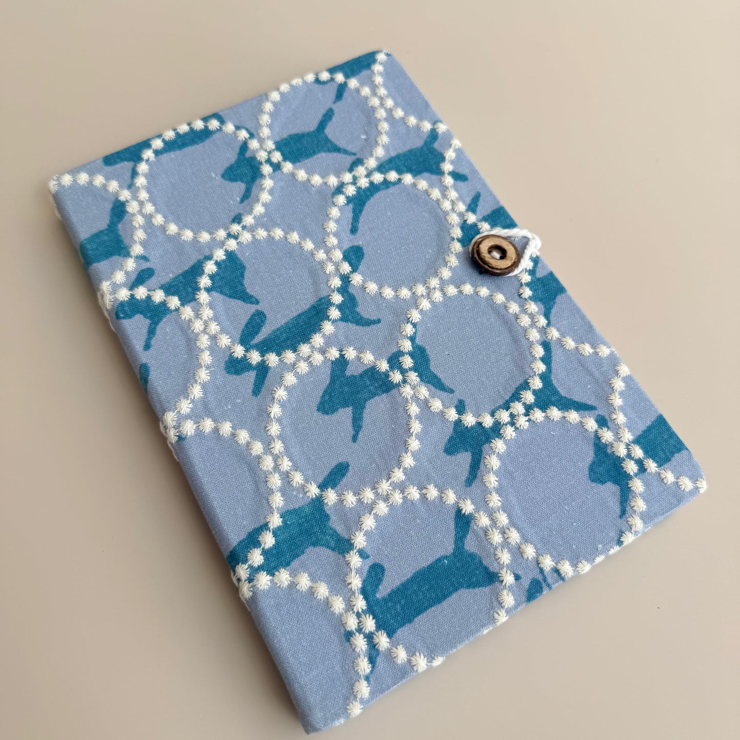 A small fabric-covered notebook or card holder with a blue and teal dachshund pattern inside white circular loops outlined with white beads, secured by a metal snap button closure, shown on a neutral beige background.