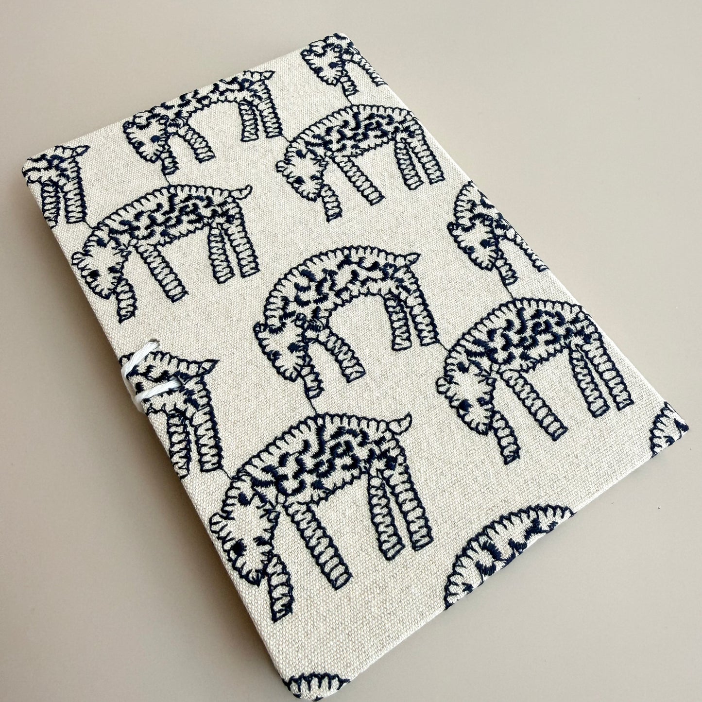 A square fabric item, likely a small notebook, journal, or pouch, featuring a navy blue and cream block-printed animal pattern on a textured linen-like fabric, photographed on a neutral beige background.