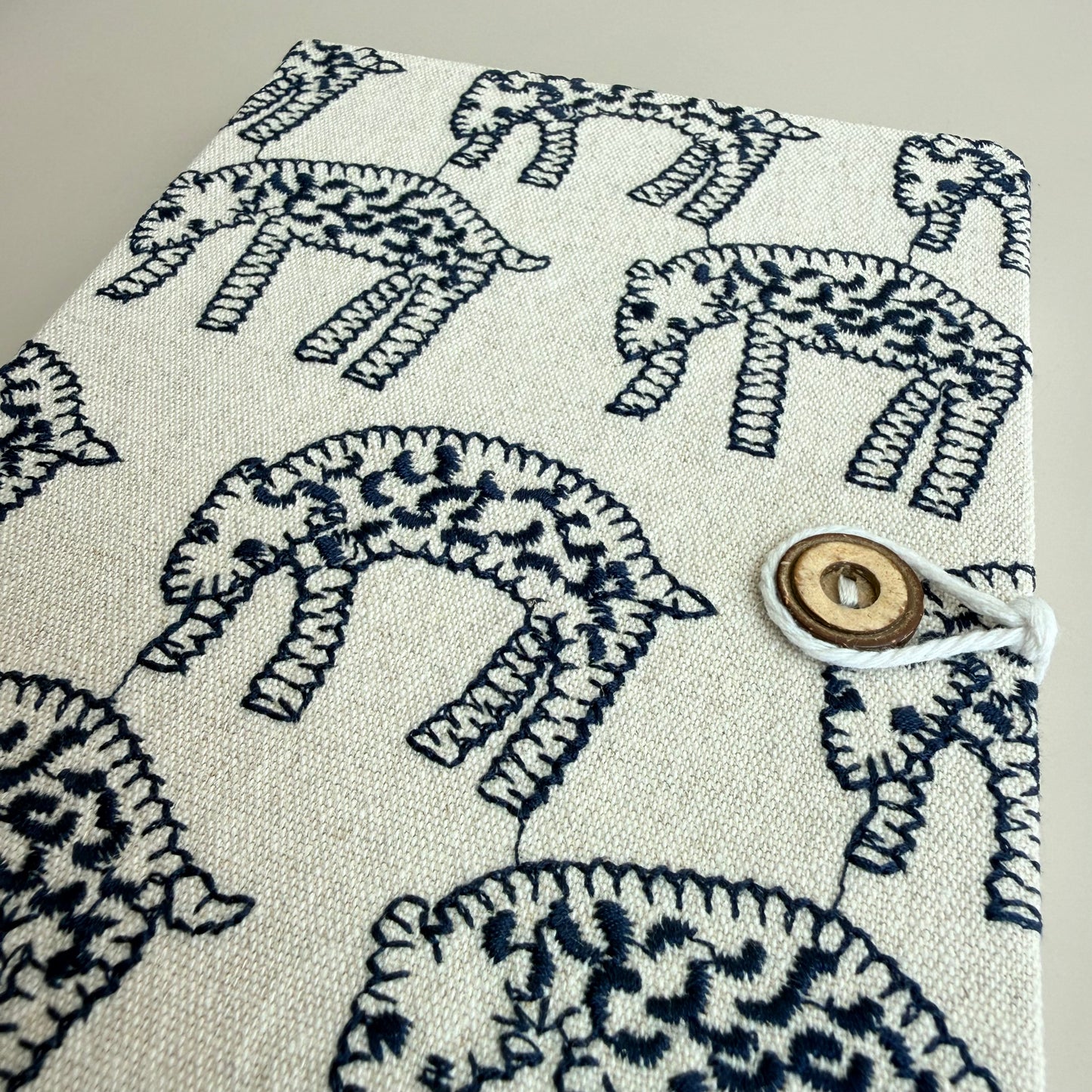 A close-up of a fabric-covered notebook or journal with a cream-colored linen-like cover featuring a dark navy blue hand-drawn style animal print. The cover is secured with a white cord and a round wooden toggle button on the right side.