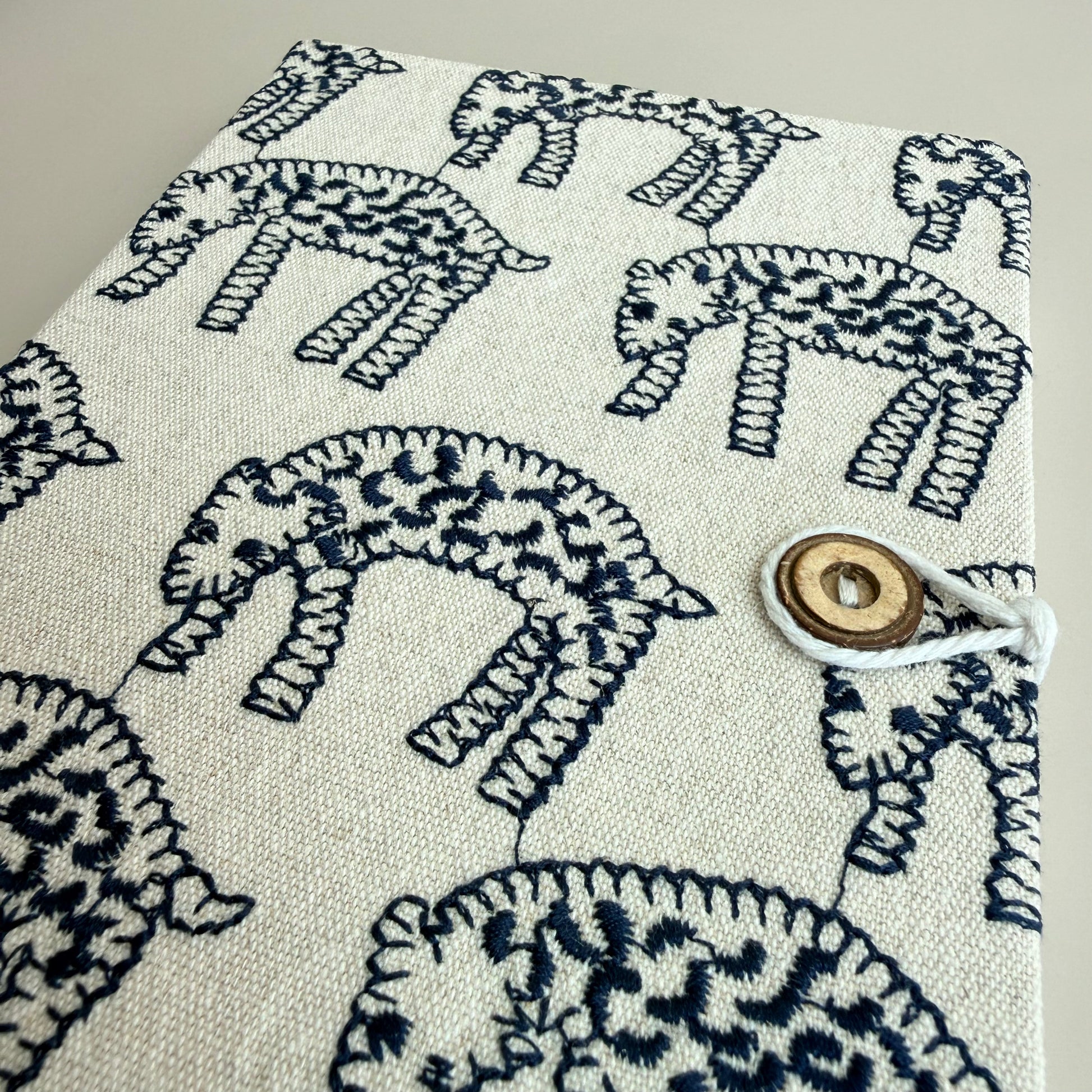 A close-up of a fabric-covered notebook or journal with a cream-colored linen-like cover featuring a dark navy blue hand-drawn style animal print. The cover is secured with a white cord and a round wooden toggle button on the right side.