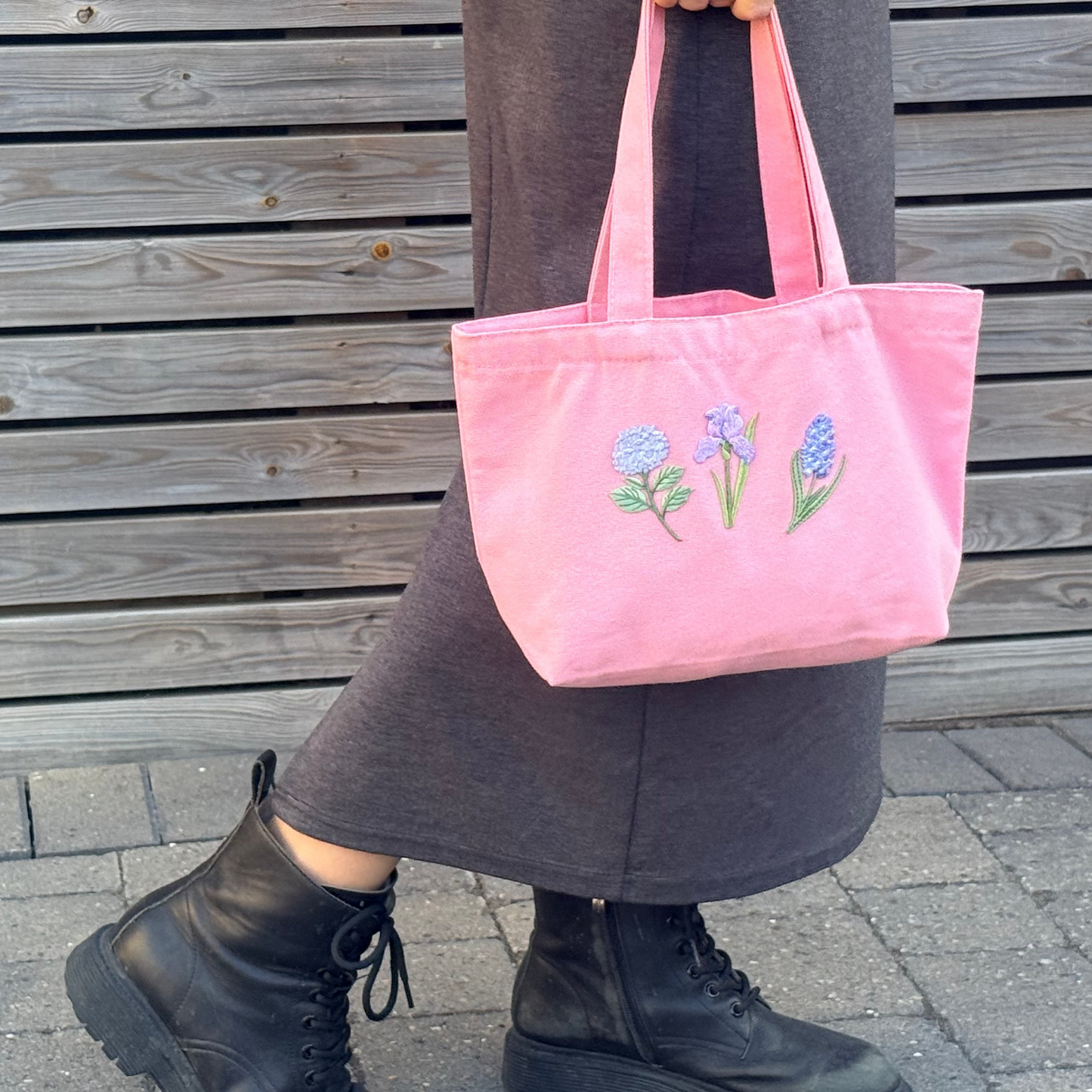 A pink canvas tote bag with embroidered blue and purple flowers, held by a person wearing a dark gray skirt and black combat boots in front of a wooden slat wall.