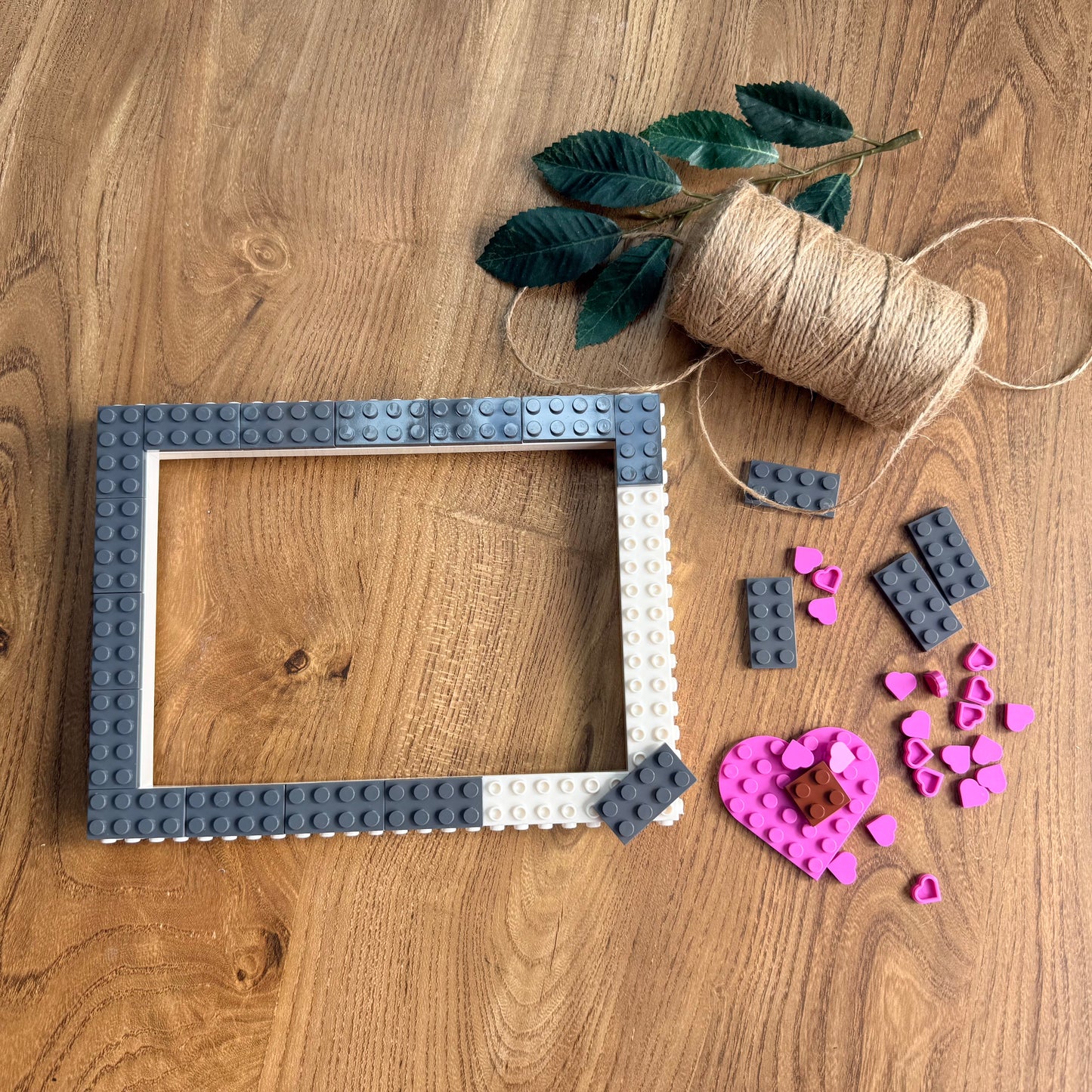 A square picture frame made from interlocking building blocks sits on a wooden tabletop, surrounded by craft supplies including a spool of twine, green leaves, loose gray blocks, and pink heart-shaped pieces.