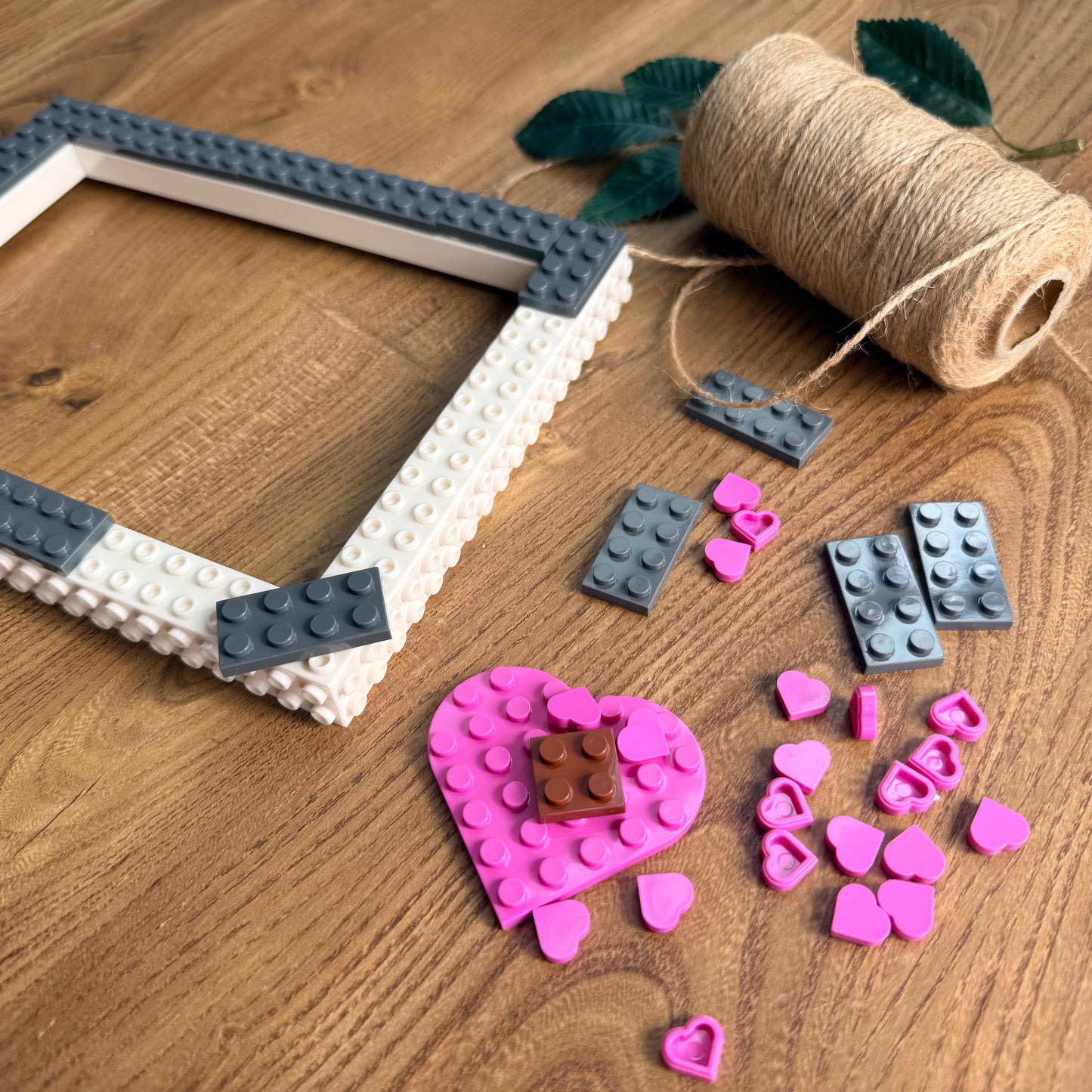 A handmade rectangular picture frame made from white and dark gray building bricks, displayed on a wooden tabletop alongside pink heart-shaped studs, loose studs, a brown brick, and a spool of twine.