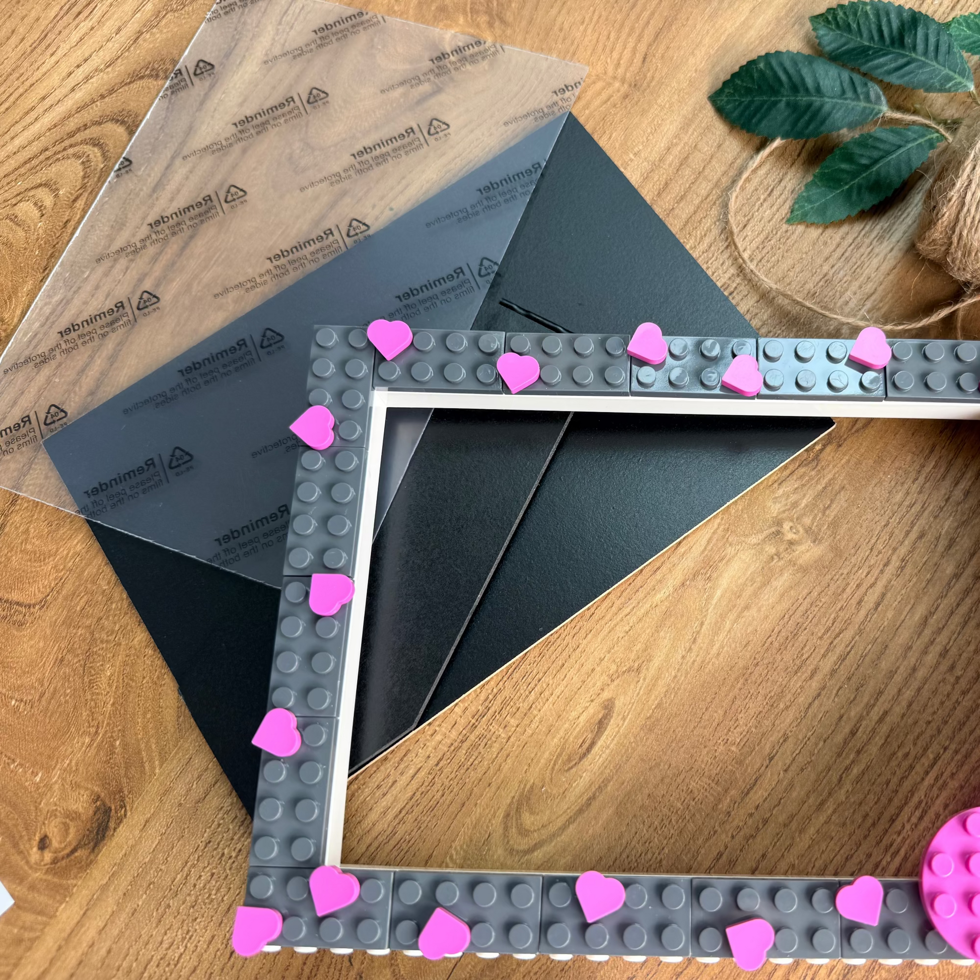 A square picture frame with a gray brick-studded border and bright pink heart accents, shown on a wooden tabletop with craft supplies including a clear sheet, black cardstock, twine, and green leaves.