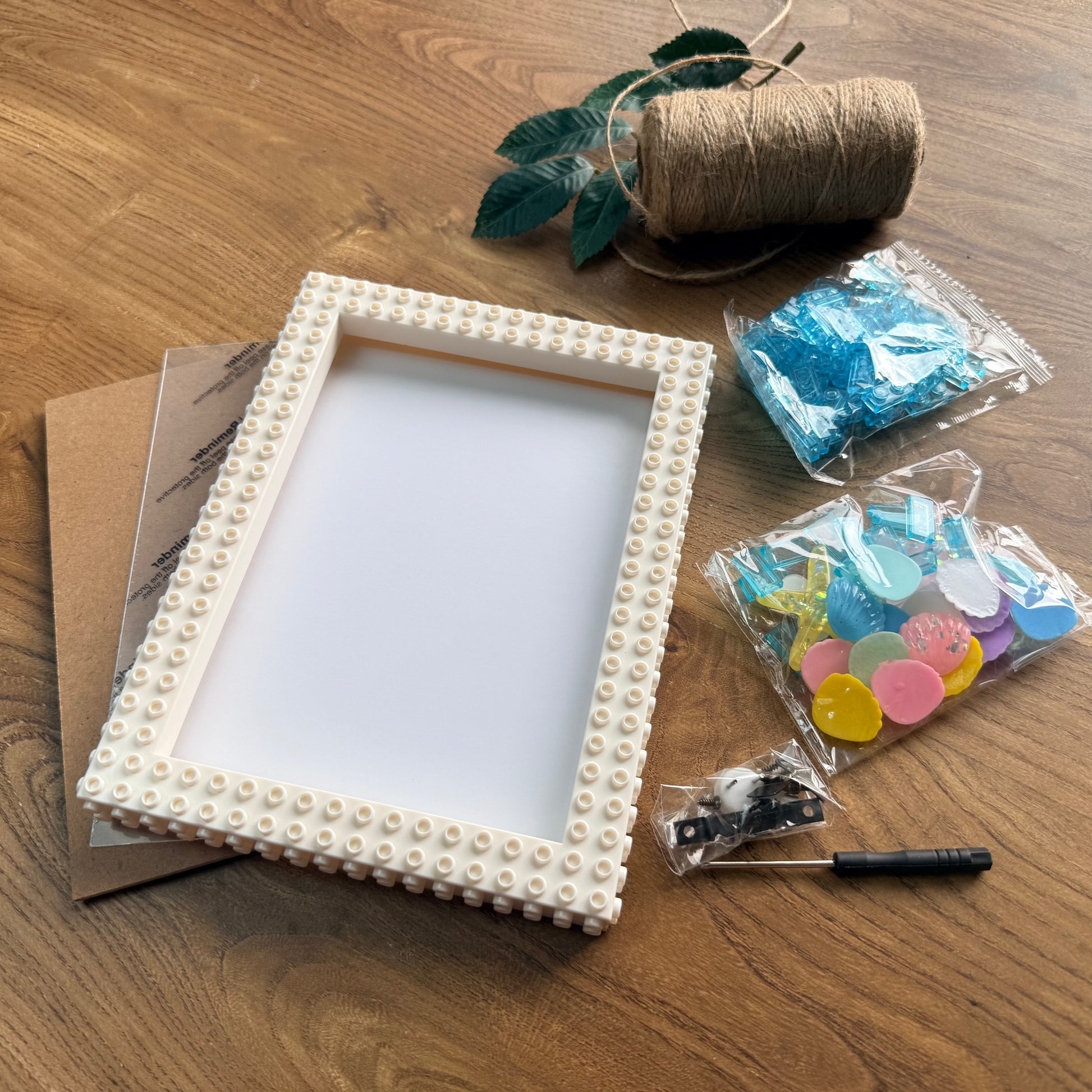 A DIY craft kit for making a pearl picture frame, displayed on a wooden tabletop. The kit includes a cream-colored wooden frame studded with small pearls, a spool of twine with green leaves, colorful round craft beads, a small screwdriver, and a backing card.