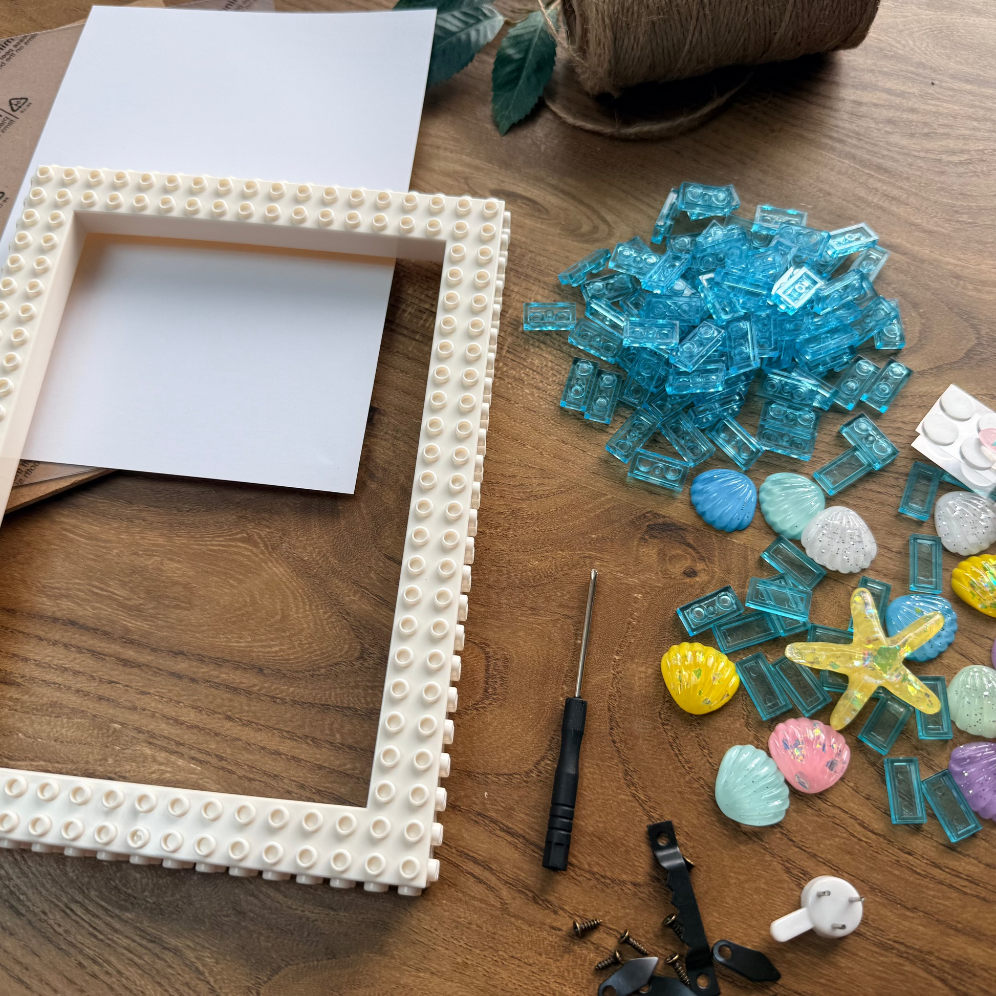 A DIY craft kit for making a pearl frame, laid out on a wooden floor. The kit includes a wooden frame studded with small white pearls, a pile of blue glass mosaic tiles, pastel seashells, a yellow starfish, small white pearls, a screwdriver, and other small decorative pieces.
