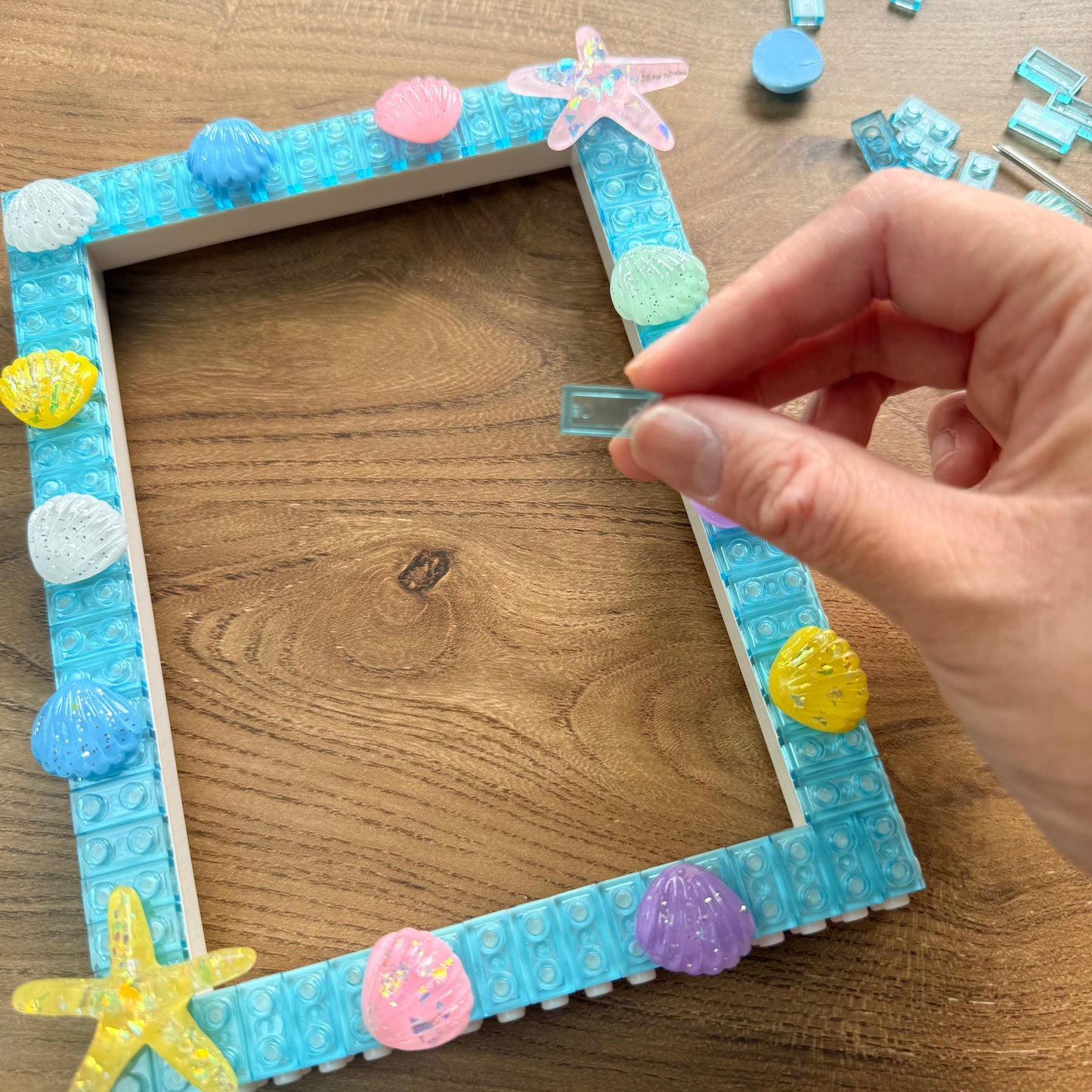 A hand is placing a small translucent blue tile into a square picture frame made of light blue building bricks, decorated with colorful seashell and starfish embellishments, on a wooden tabletop.