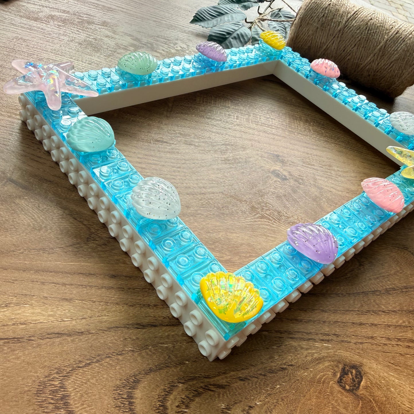 A square white picture frame decorated with translucent blue studs, colorful seashell and starfish embellishments, displayed on a wooden tabletop.