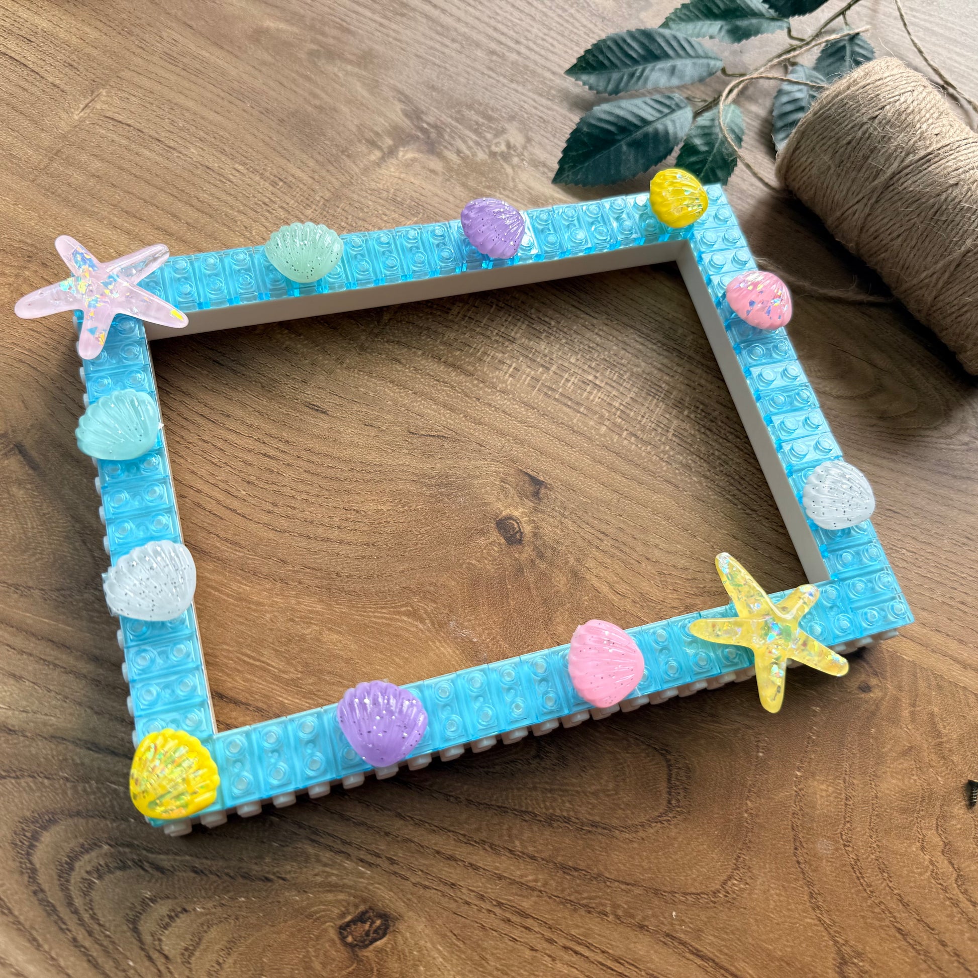 A square decorative picture frame made from light blue LEGO bricks, embellished with colorful seashell and starfish charms, displayed on a wooden tabletop.