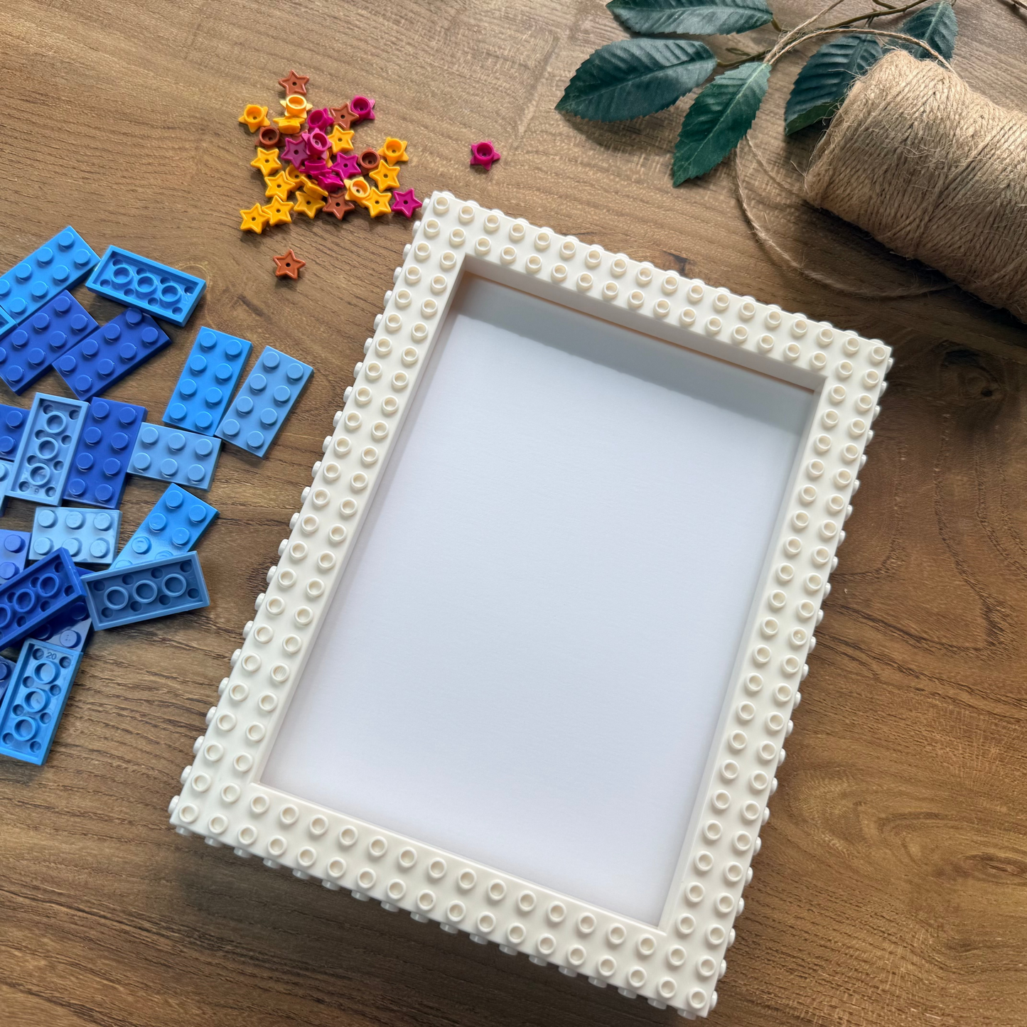 An off-white rectangular picture frame with a textured, Lego-like raised stud border, displayed on a wooden tabletop alongside blue building blocks, colorful star-shaped embellishments, green leaves, and a spool of twine.