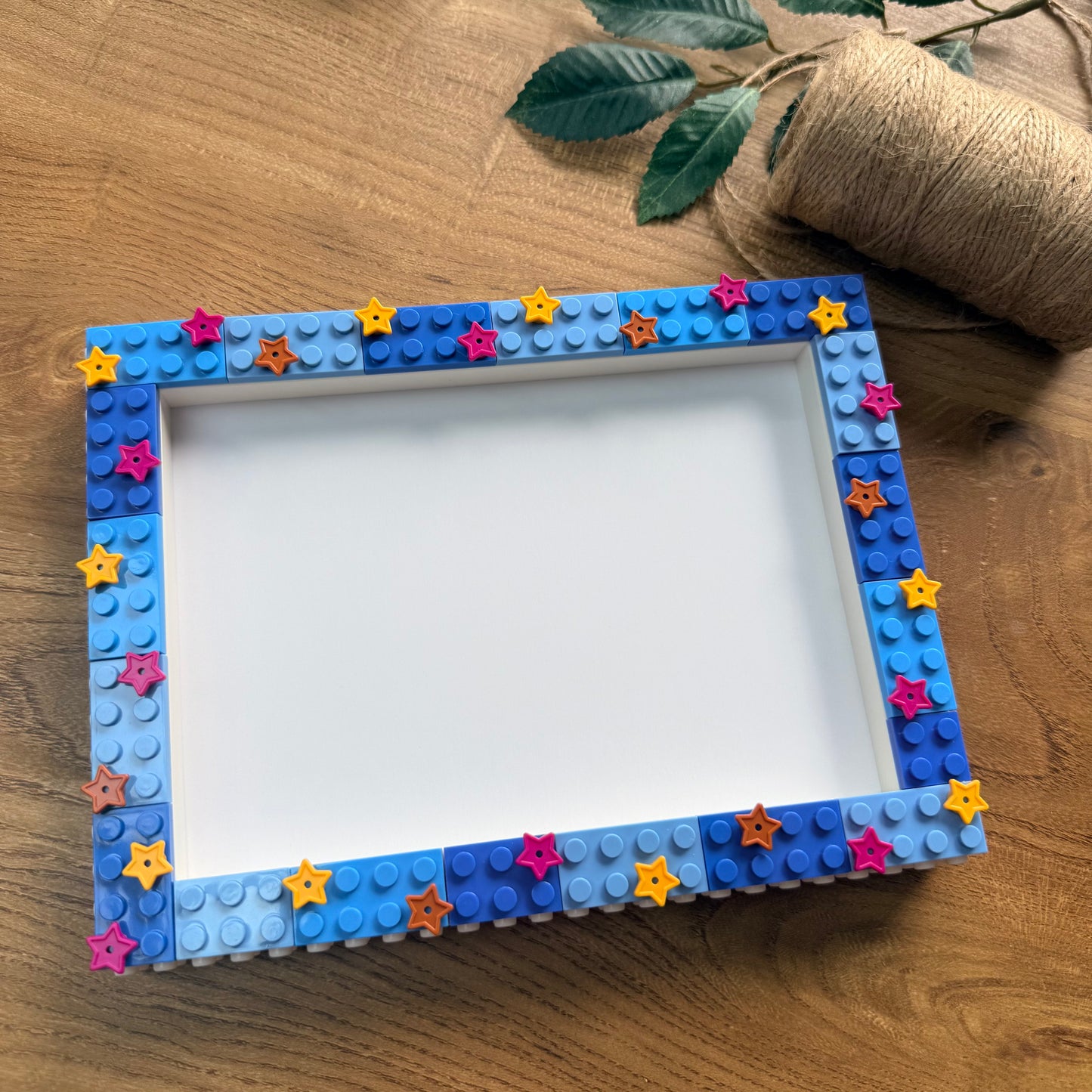 A square picture frame decorated with blue and light blue toy building-block studs and small colorful star stickers, placed on a wooden surface with a twine roll and green leaves in the background.