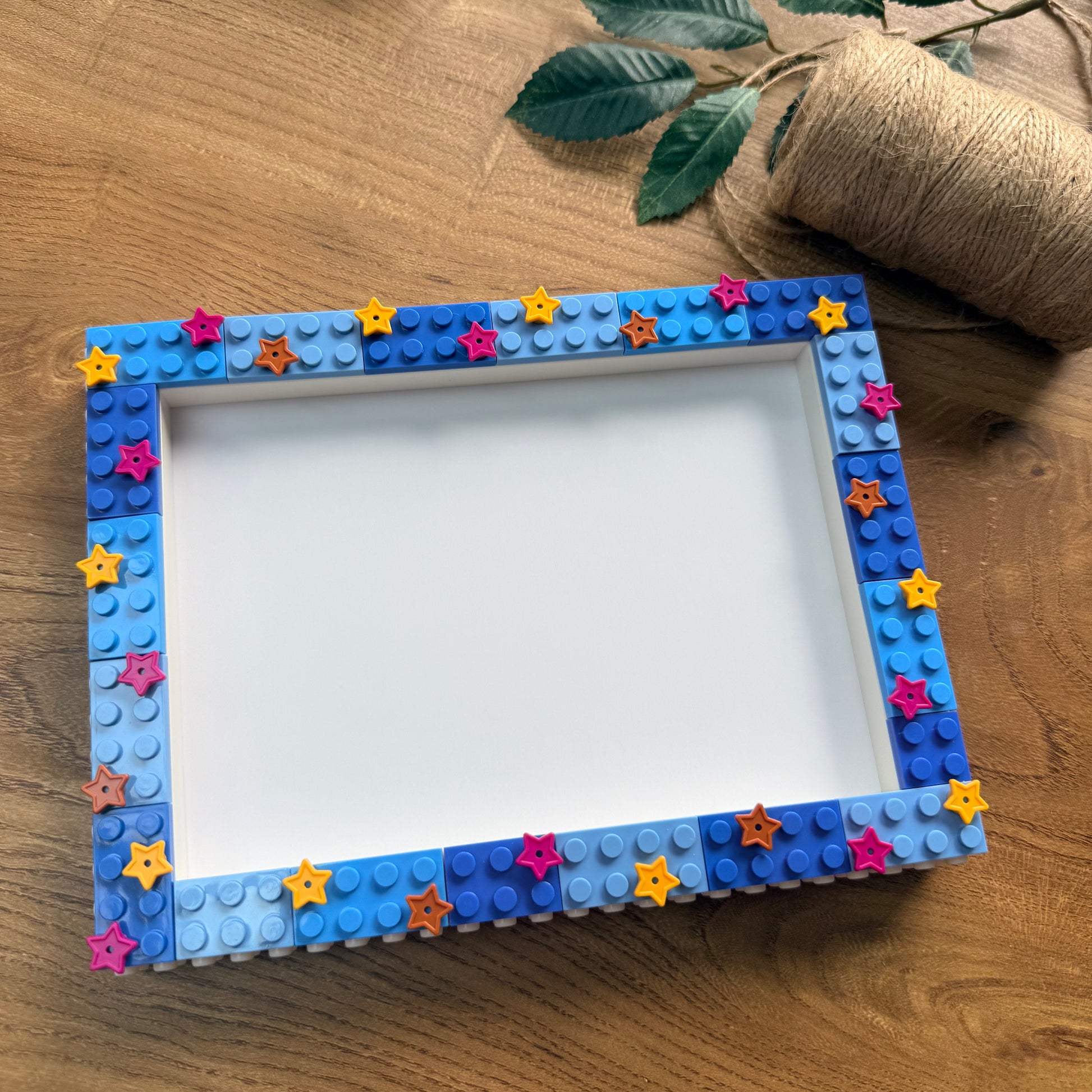 A square picture frame decorated with blue and light blue toy building-block studs and small colorful star stickers, placed on a wooden surface with a twine roll and green leaves in the background.
