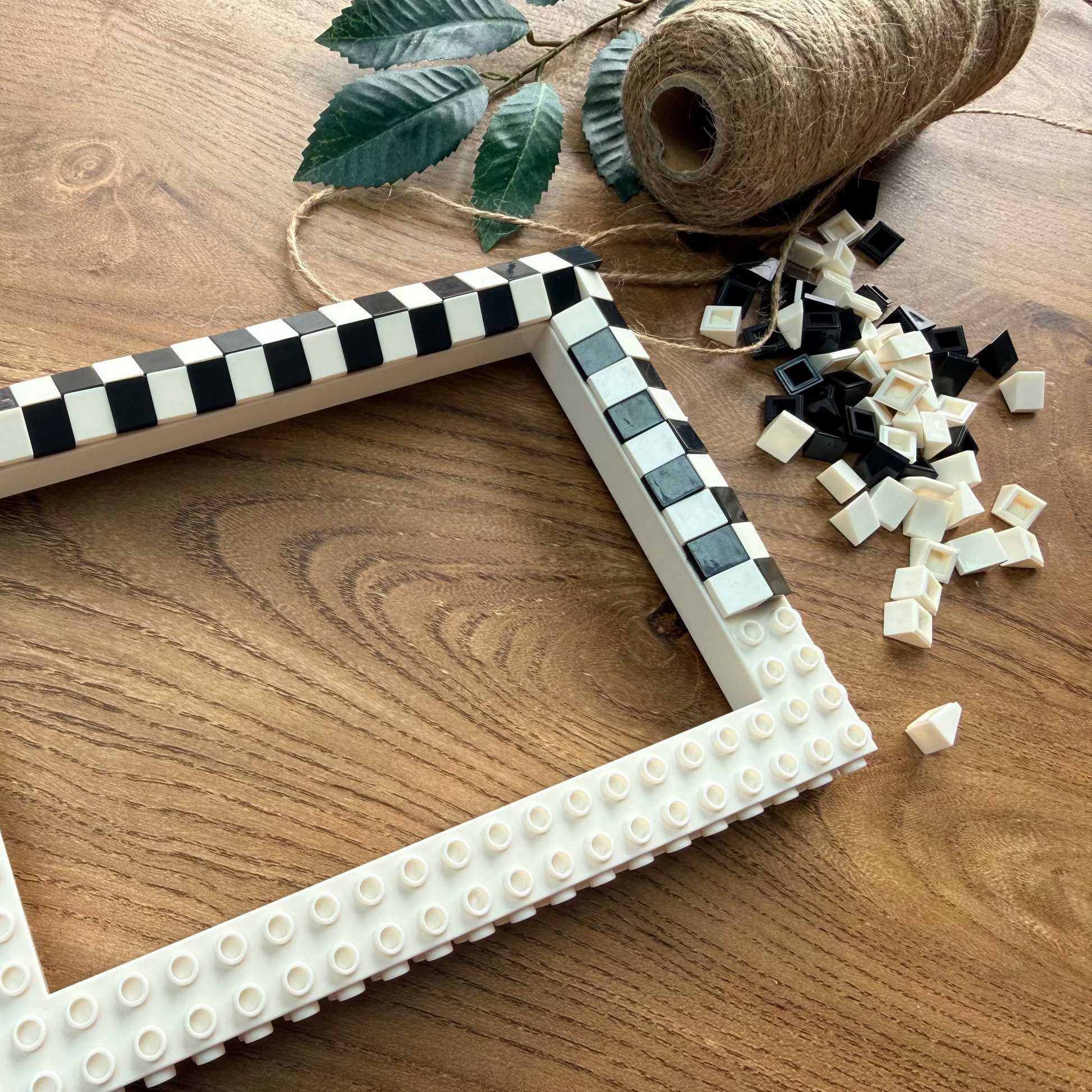 A rectangular picture frame made from white and black checkered tiles, styled on a wooden tabletop with craft scraps, twine, and leaves nearby.