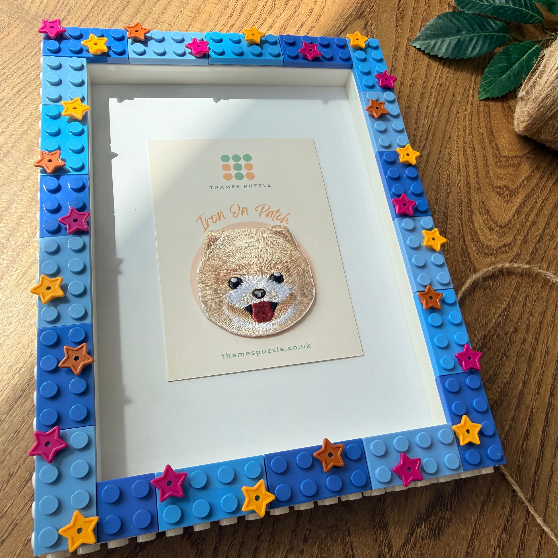 A custom framed picture made from blue building blocks with colorful star accents, displaying an embroidered dog portrait. The frame is styled on a wooden tabletop next to a spool of twine.